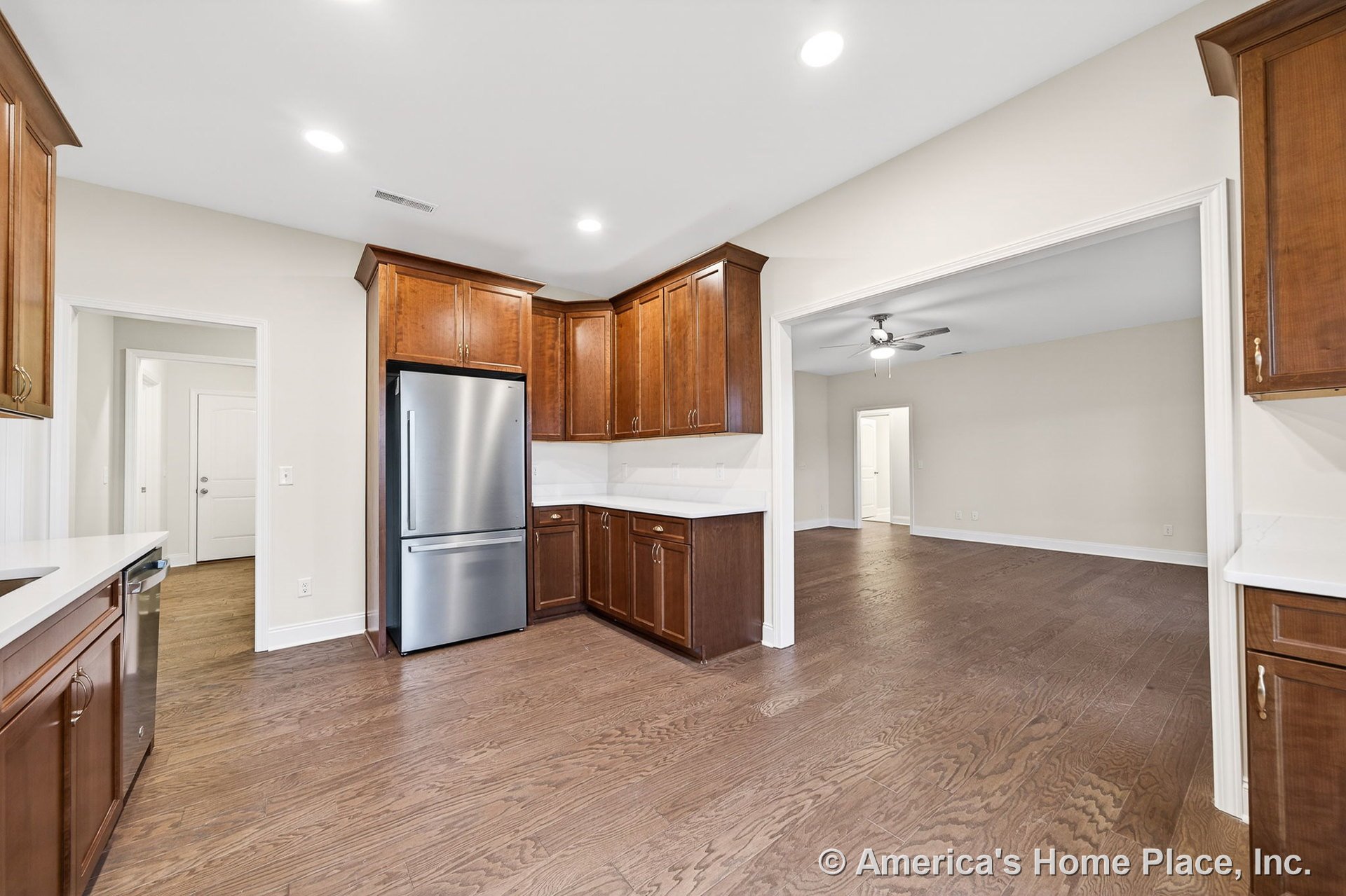 Wood kitchen cabinetry with quartz countertops and stainless steel refrigerator, recessed ceiling lights, hardwood flooring extending into open living room with ceiling fan and