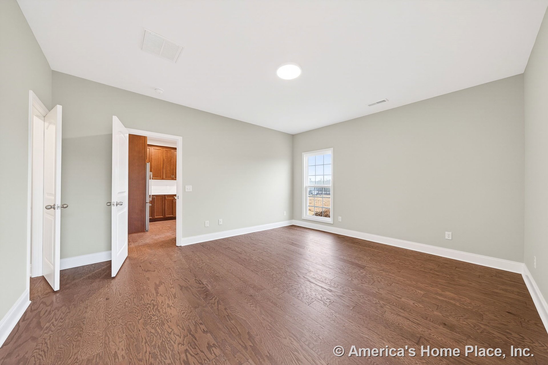Hardwood flooring and neutral walls with white trim, large window providing natural light, double doors opening to kitchen with wood cabinetry, recessed ceiling light, baseboards