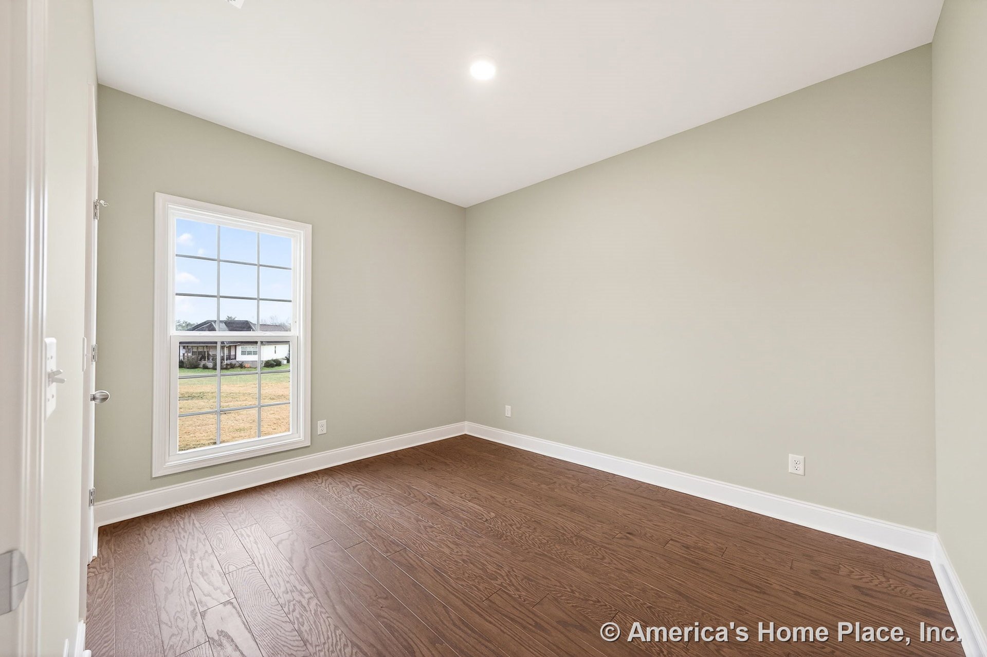 Bedroom with light green painted walls, white baseboards and trim, wood plank flooring, large single window with white casing, recessed ceiling light, and abundant natural light in