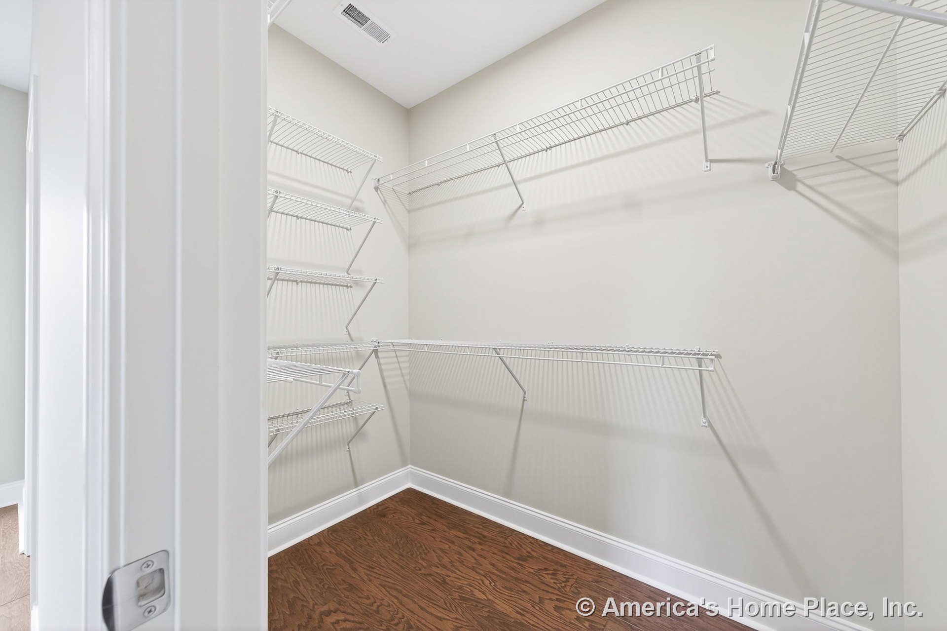 Walk-in closet with multiple white wire shelving units, hardwood flooring, neutral wall color, white baseboards, ceiling vent, and door frame trim.