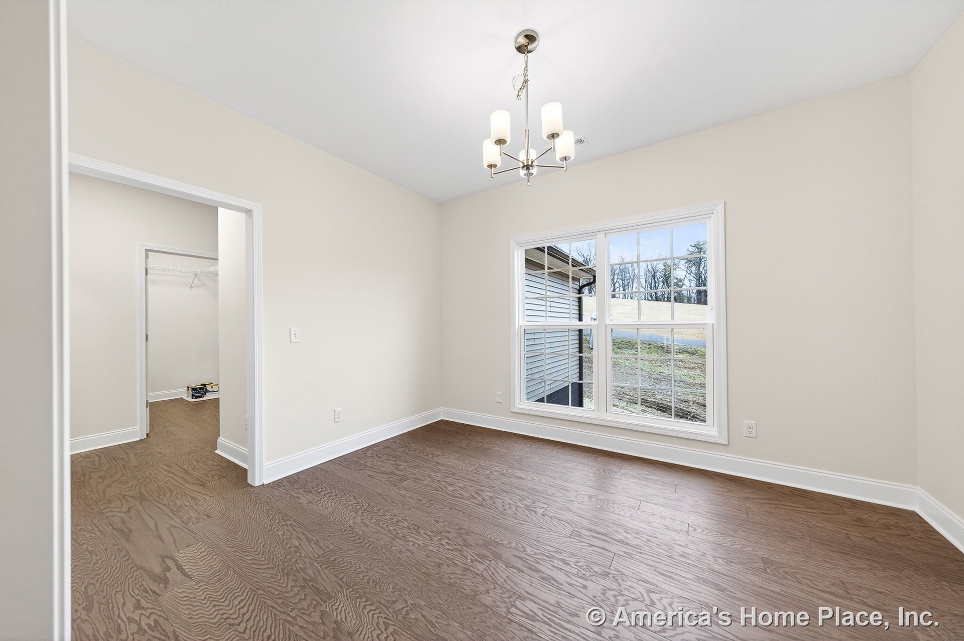 Dining room with wide plank wood flooring, large window with white trim and divided lights, modern ceiling chandelier, neutral wall color, white baseboards, and open doorway