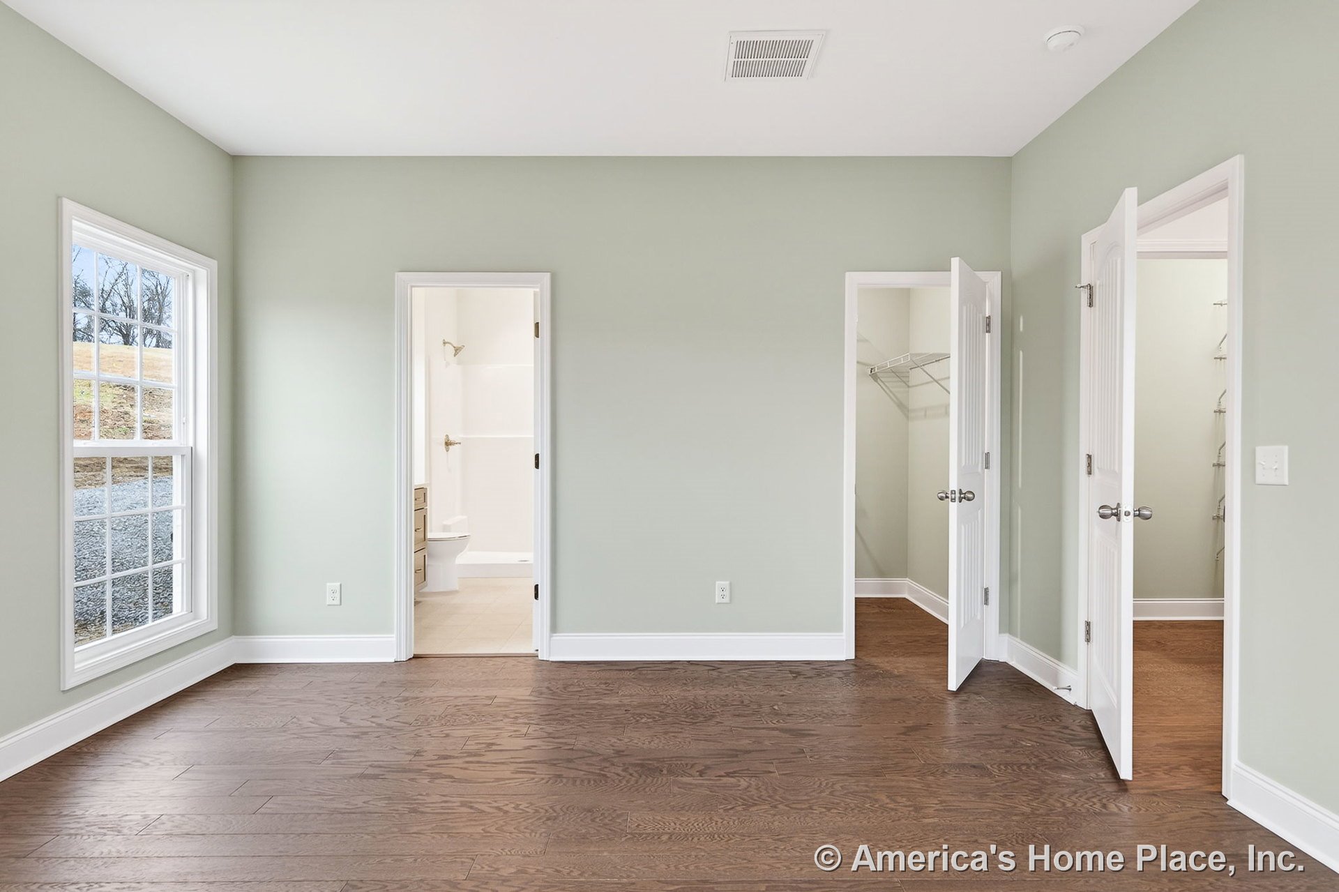 Bedroom with hardwood flooring, large window framed by white trim, light green painted walls, white paneled doors leading to a walk-in closet with wire shelving and a bathroom
