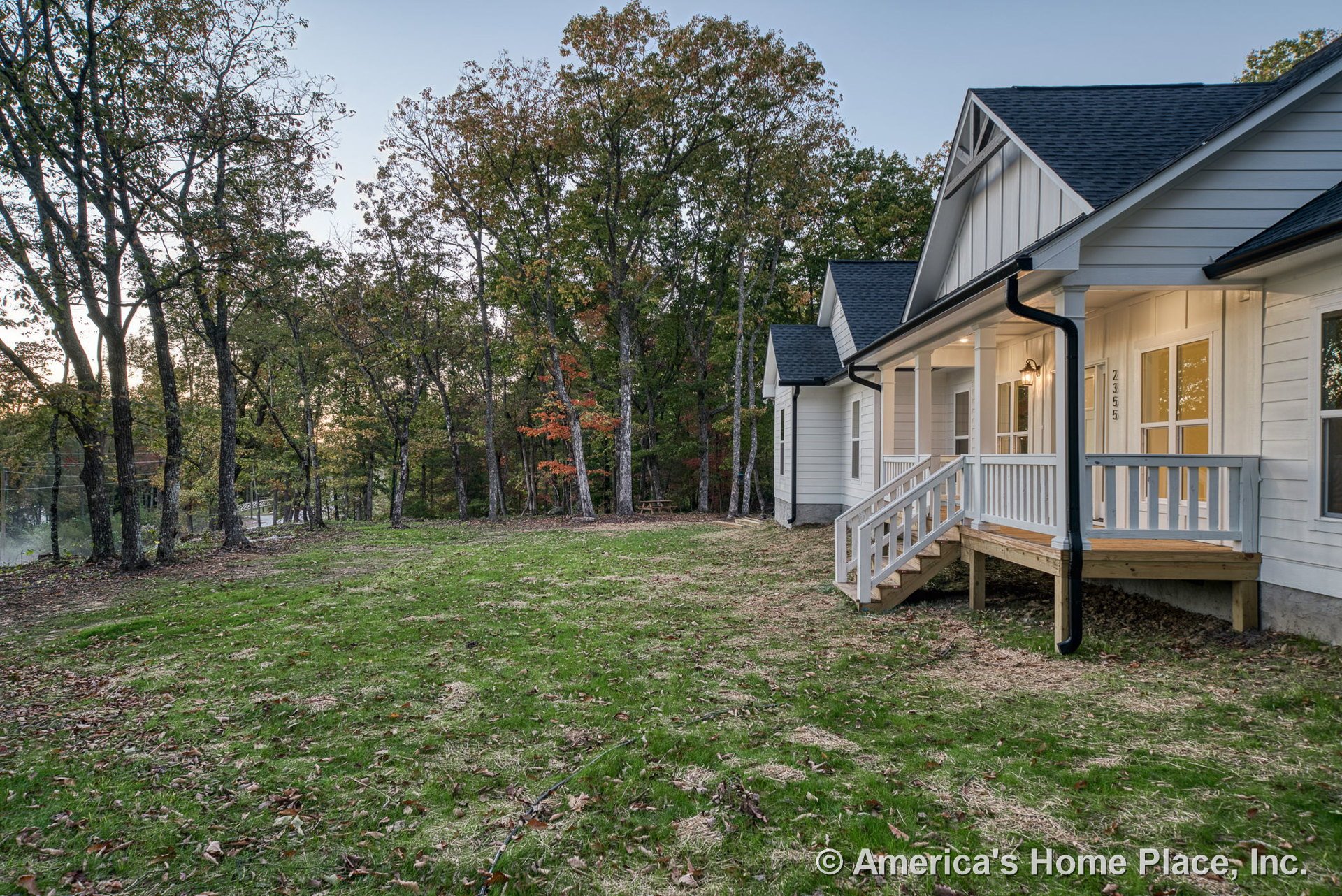 Covered front porch with white railings and wooden steps, white exterior siding, black shingle roof, multiple windows with white trim, exterior lighting fixture near entry door