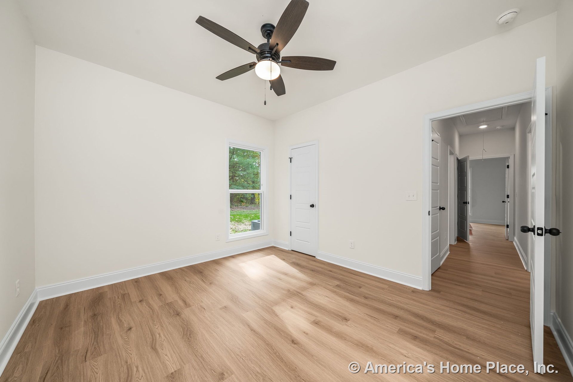 Bedroom with light wood plank flooring, white trim and baseboards, single window offering outdoor view, ceiling fan with integrated light, white paneled doors, neutral wall color