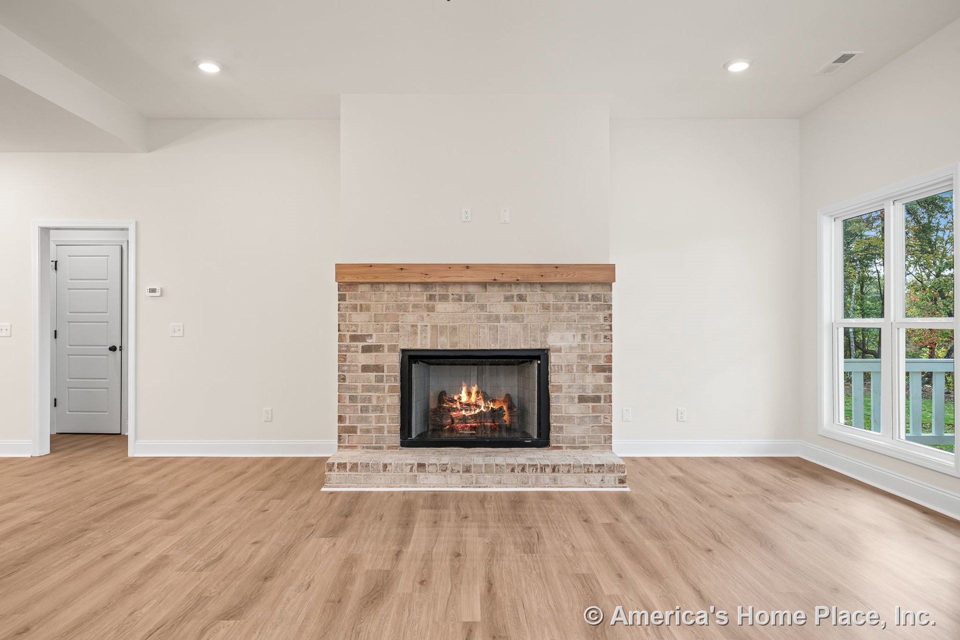 Brick fireplace with wood mantel centered on white wall, flanked by large windows; light wood flooring, white trim, recessed ceiling lights, and interior door visible.