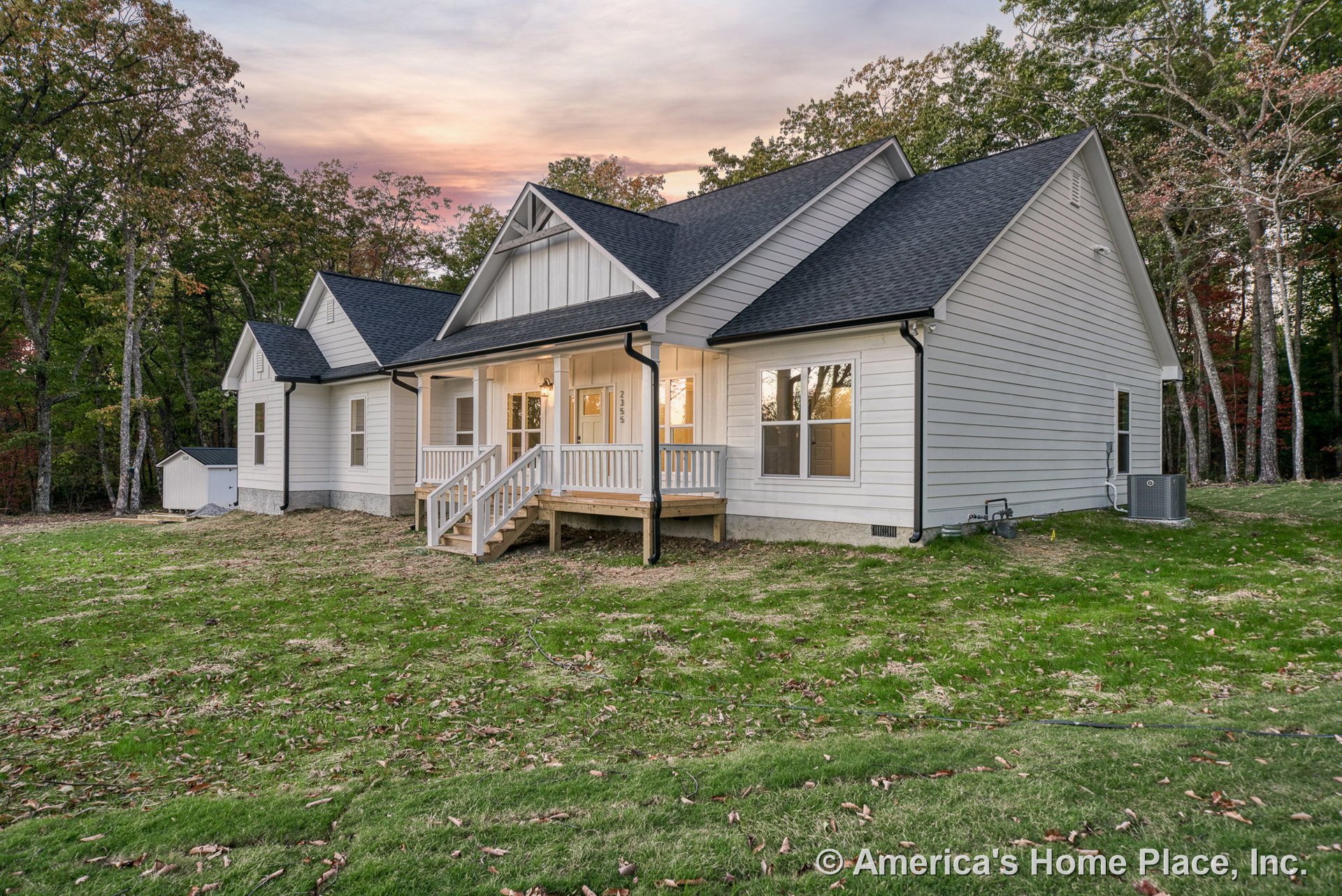 Covered front porch with white railing, board-and-batten siding on the gable, multiple double-hung windows, black shingle roof, exterior lighting fixture, and white trim around the