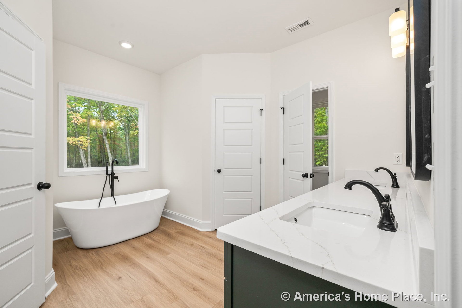 Freestanding soaking tub beside a large window with white trim, double sink vanity topped with quartz countertops, matte black faucets, wood-look flooring, recessed lighting