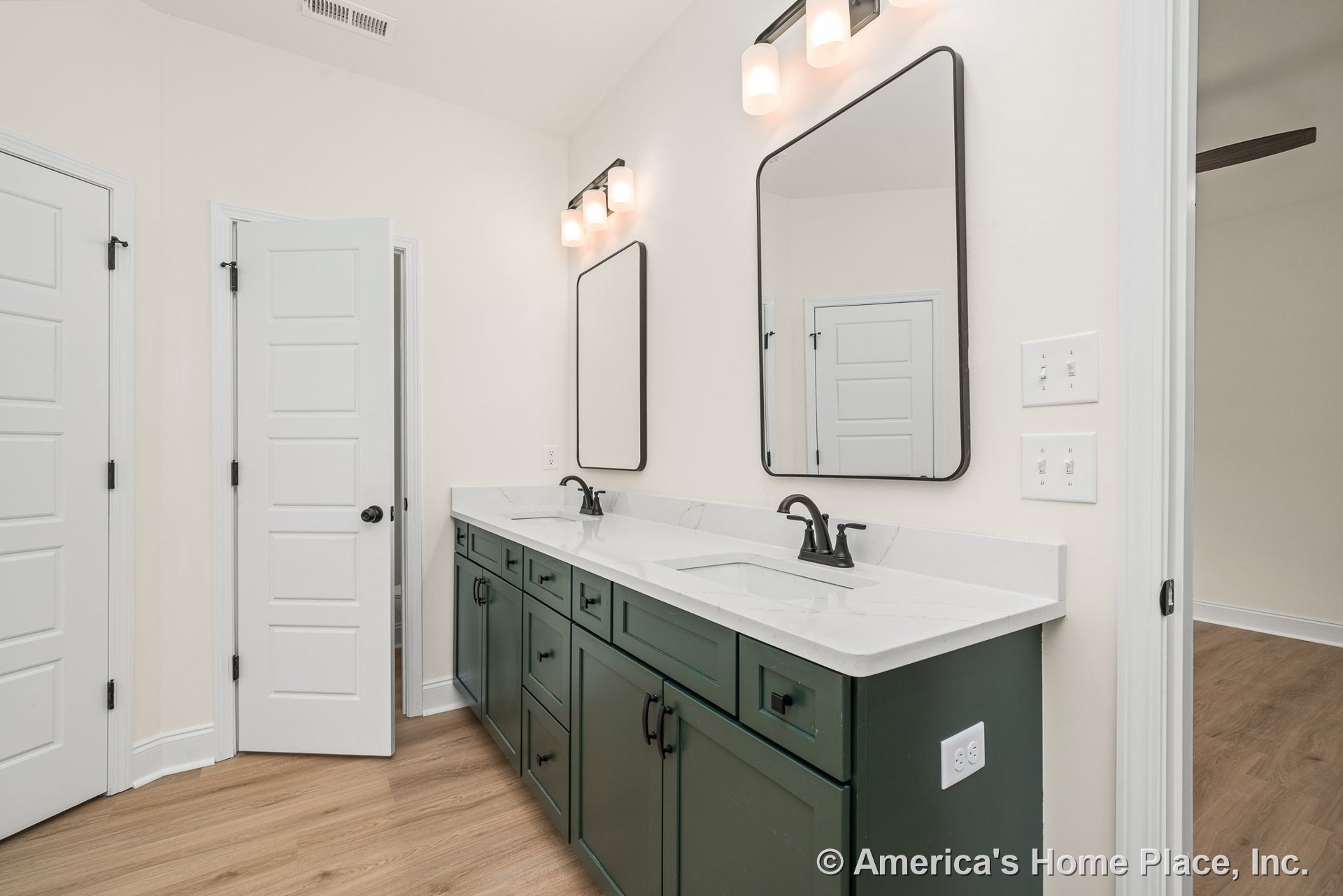 Double vanity with green shaker cabinets and white quartz countertops, wall-mounted mirrors, modern vanity lighting, matte black fixtures, wood-look flooring, white doors and trim.
