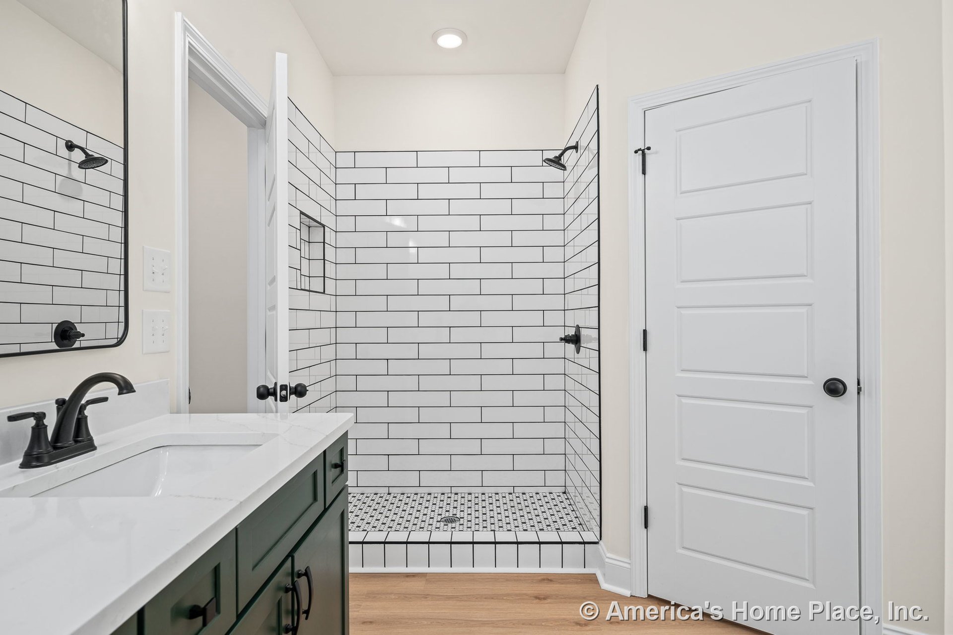 Walk-in shower with white subway tile walls and matte black fixtures, green vanity cabinetry topped with white quartz countertop, wood-look flooring, paneled interior door