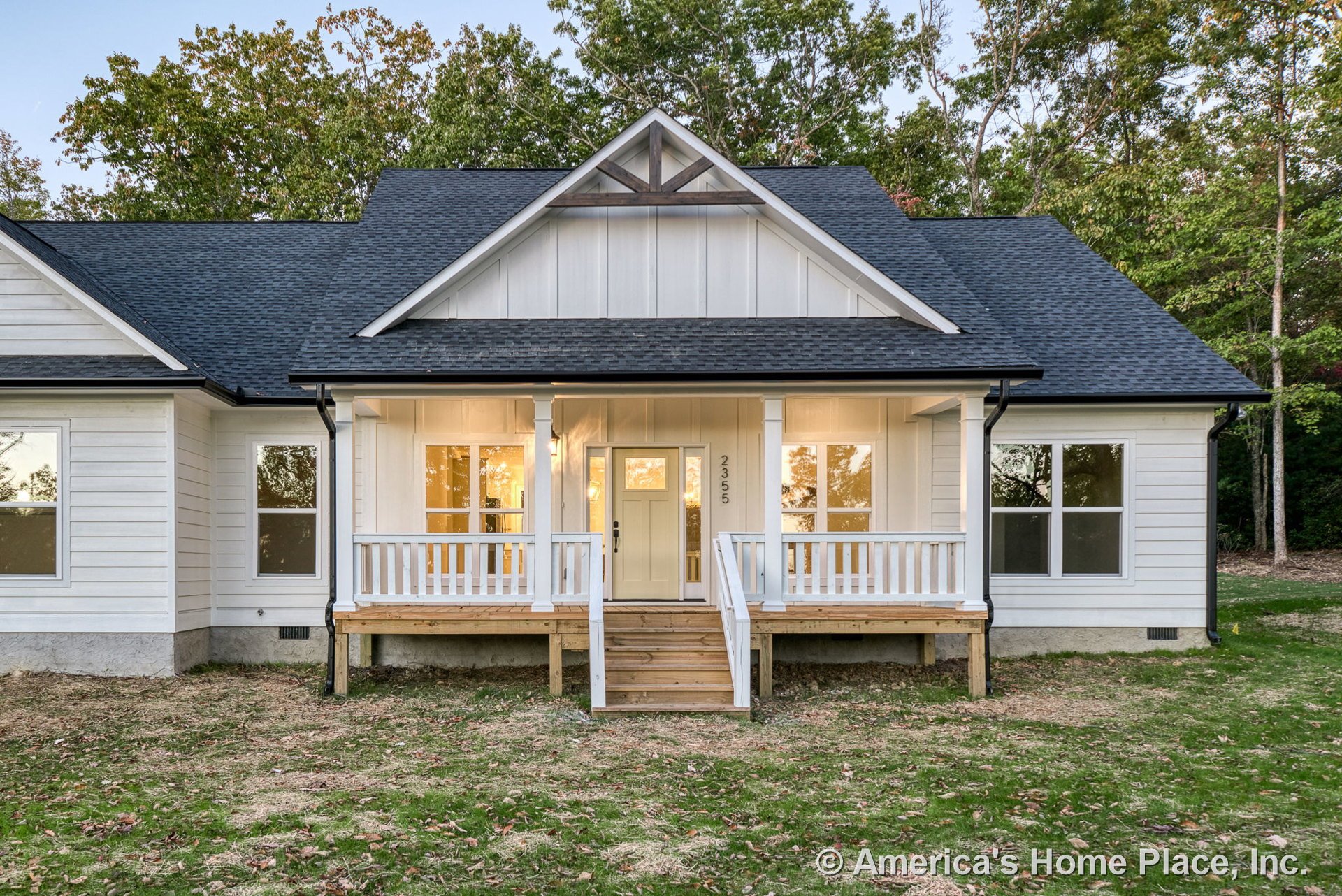 Covered front porch with white railings, board-and-batten siding, black shingle roof, double-hung windows, wooden steps, decorative gable trim, and exterior lighting.