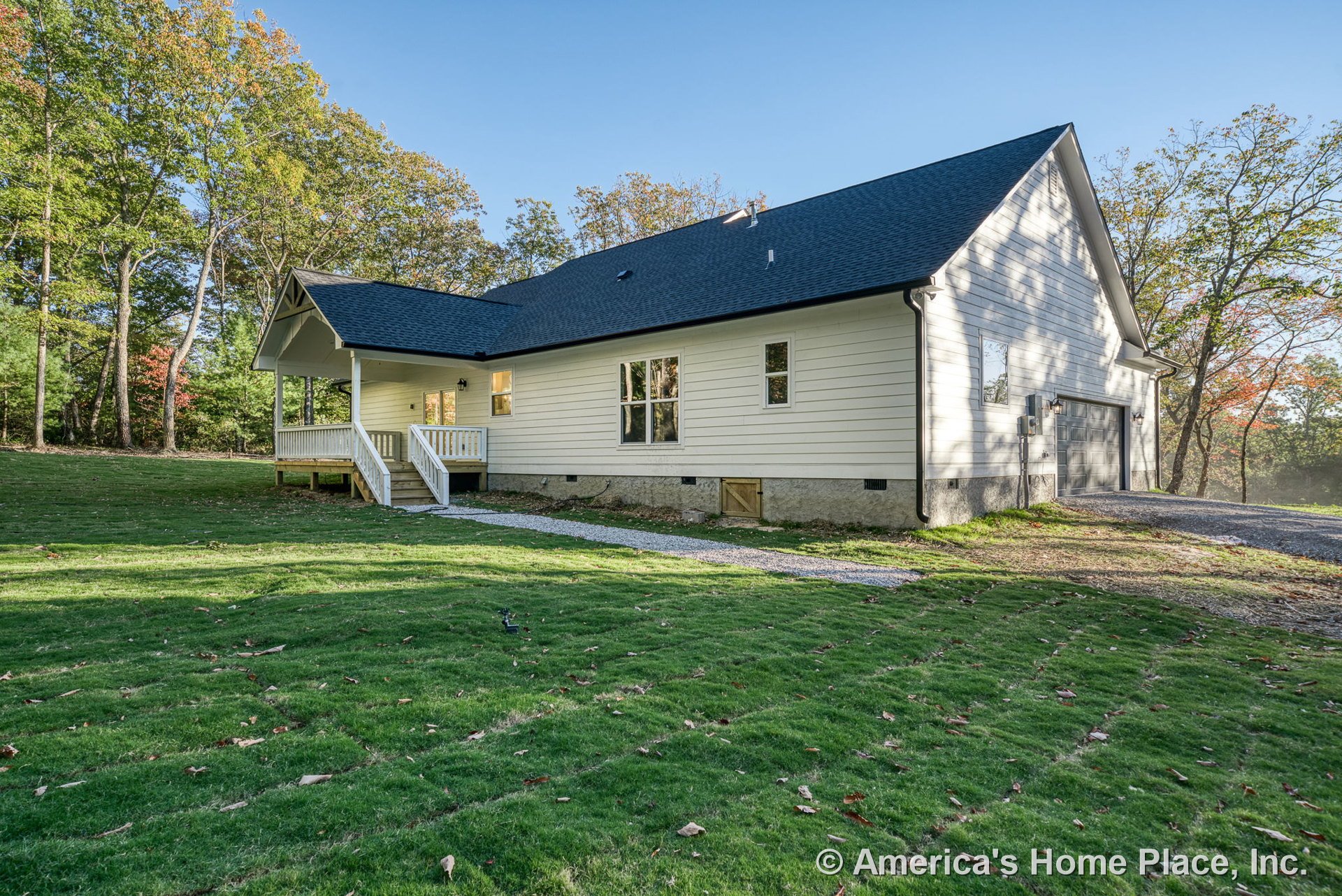 White horizontal siding and black shingle roof, covered front porch with white railings and steps, attached two-car garage, double-hung windows, gravel driveway, exterior lighting
