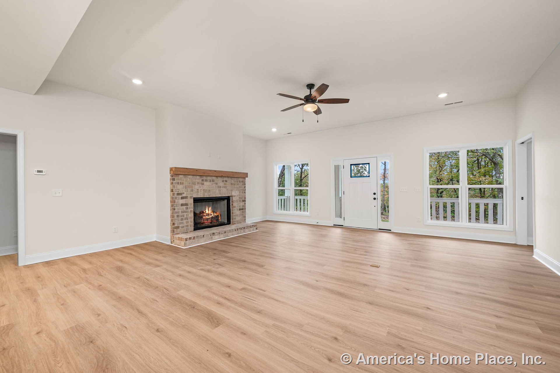 Brick fireplace with wood mantel, light wood plank flooring, large white framed windows, ceiling fan with light, recessed ceiling lights, and white front door with glass insert in