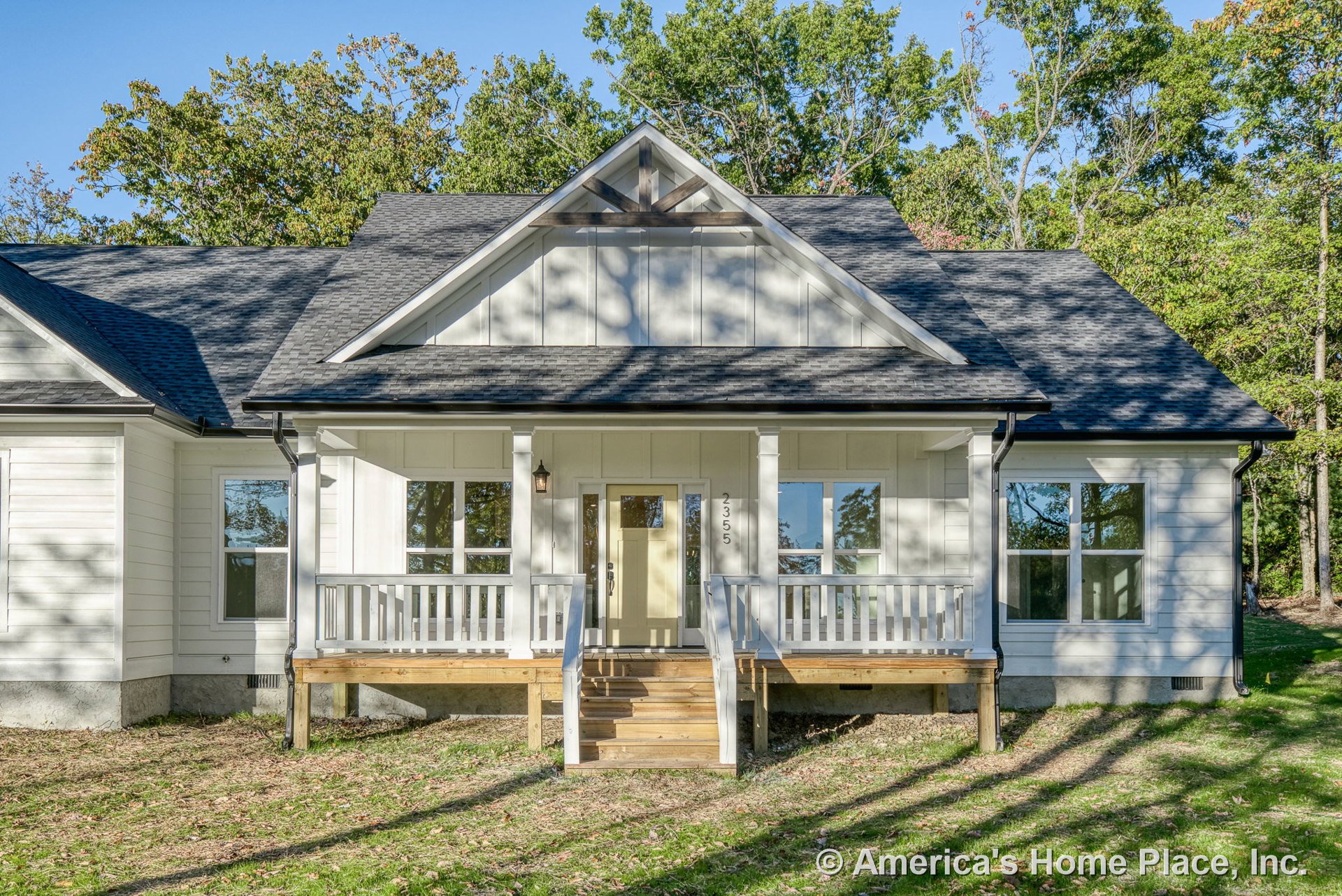 Covered front porch with white railings and columns, board-and-batten siding, large front windows, gable roof with architectural trim, wooden steps, black shingle roofing, and