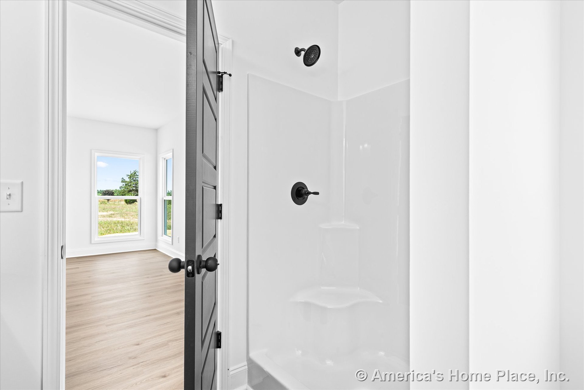 White fiberglass shower enclosure with matte black fixtures beside a paneled interior door, opening to a bright room with large picture windows, light wood plank flooring, white