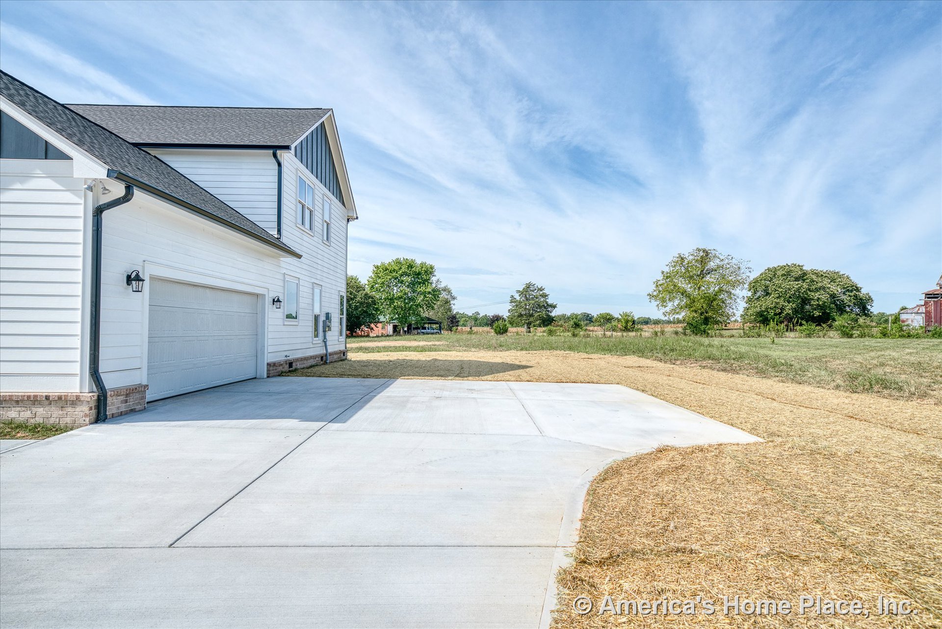 Two-car garage with white horizontal siding, concrete driveway, brick foundation trim, exterior wall-mounted lights, and multiple rectangular windows on a two-story home.