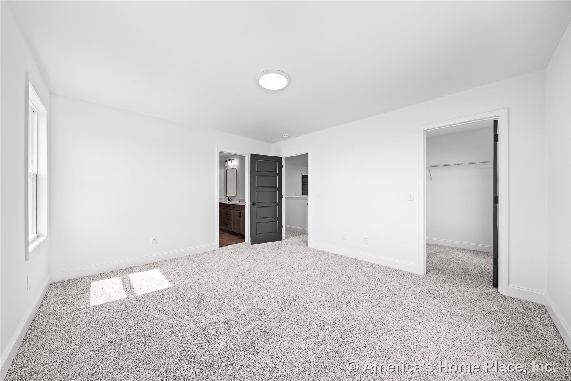Bedroom with light carpet flooring, white walls, black paneled door opening to walk-in closet with wire shelving and en suite bathroom featuring a vanity with built-in lighting