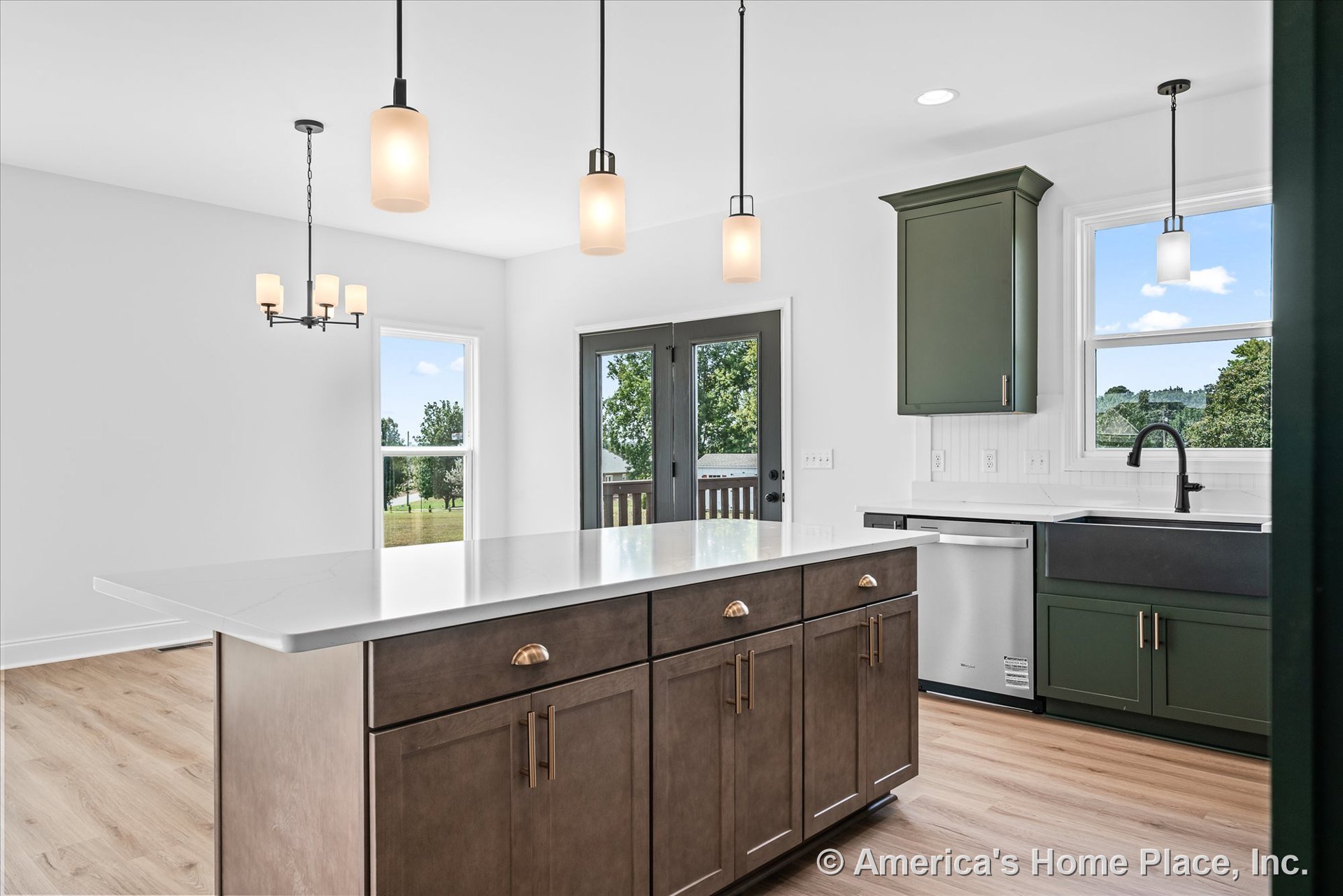 Spacious kitchen with large quartz island, green upper and lower cabinets, matte black faucet, pendant and chandelier lighting, double glass patio doors, light wood flooring, and
