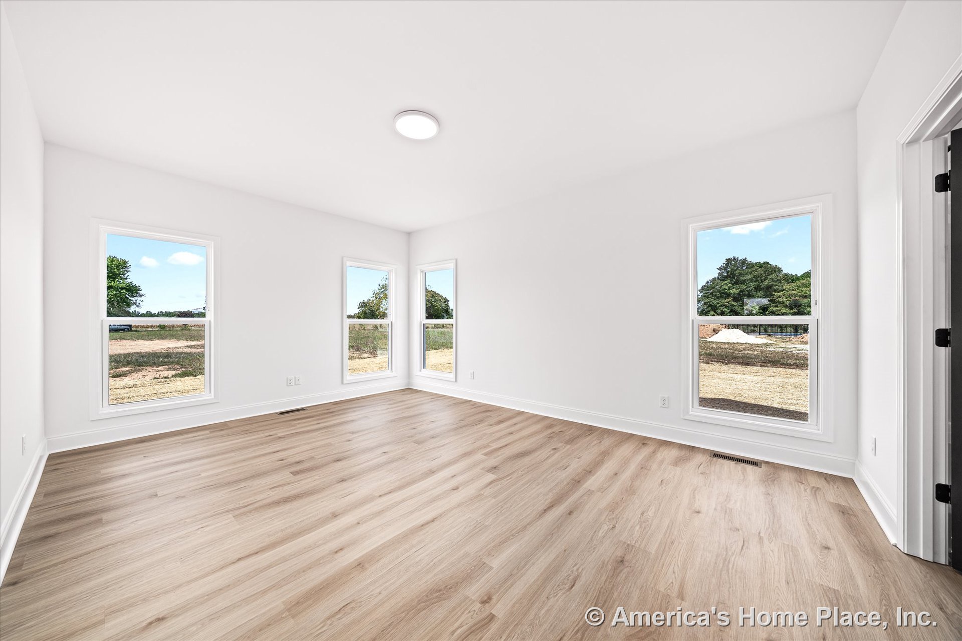 Bright interior with three large windows, light wood plank flooring, white painted trim, flush mount ceiling light, white walls, baseboard molding, and floor vent.