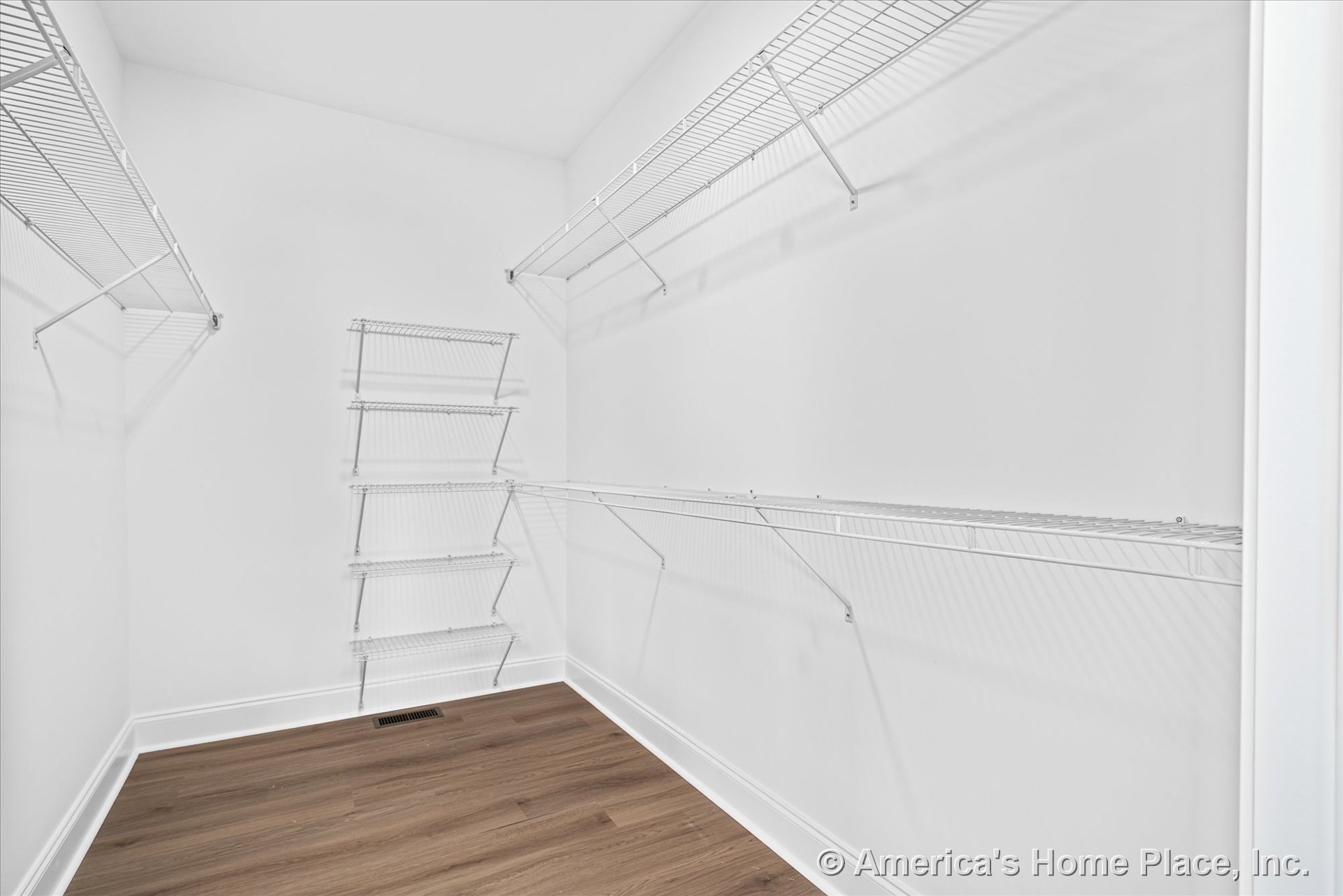 Walk-in closet with multiple white wire shelving units, wood-look flooring, white walls, baseboard trim, and a floor vent.