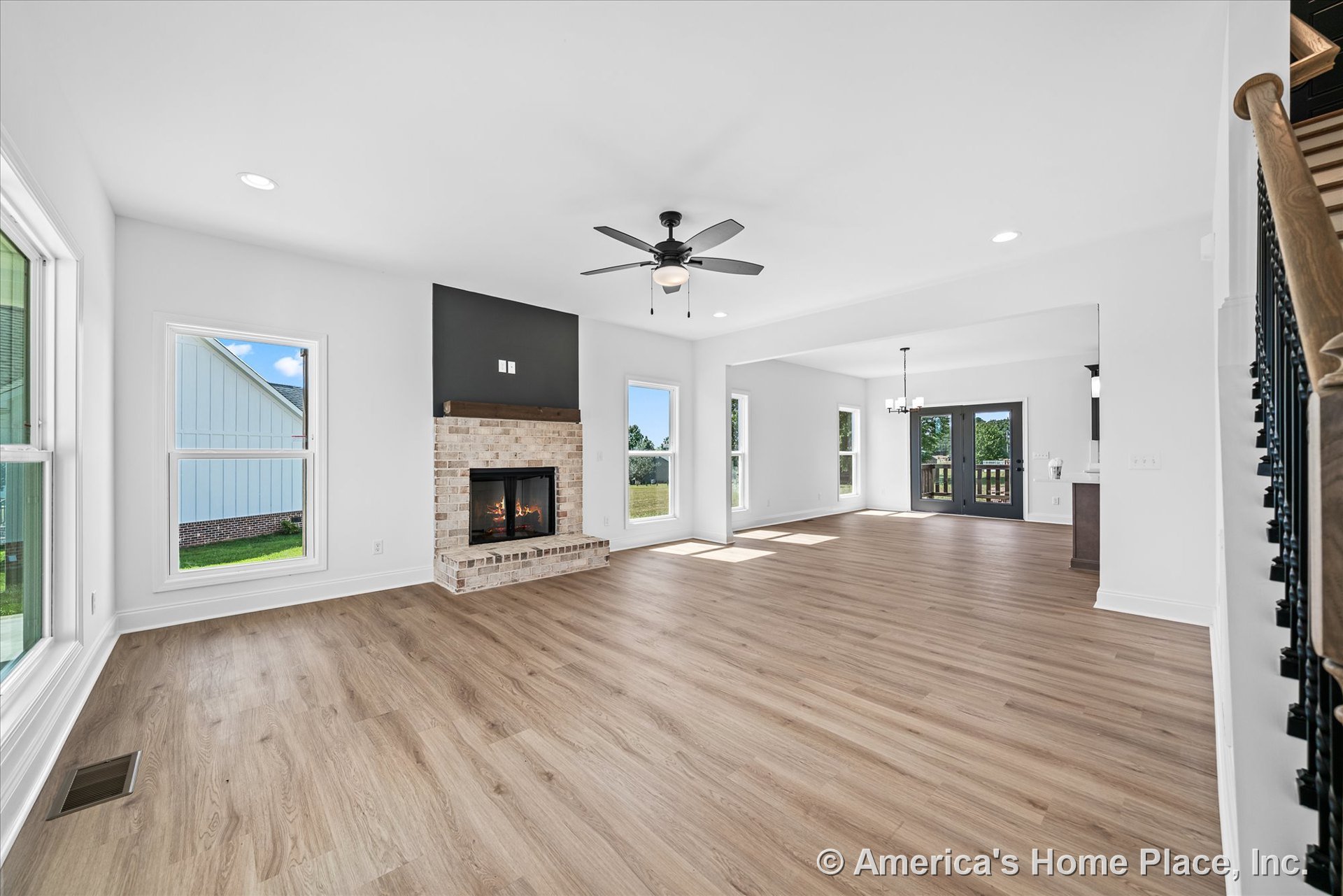 Brick fireplace with wood mantel, light wood plank flooring, large white-trimmed windows, black ceiling fan with light, open layout connecting to dining area, double glass doors