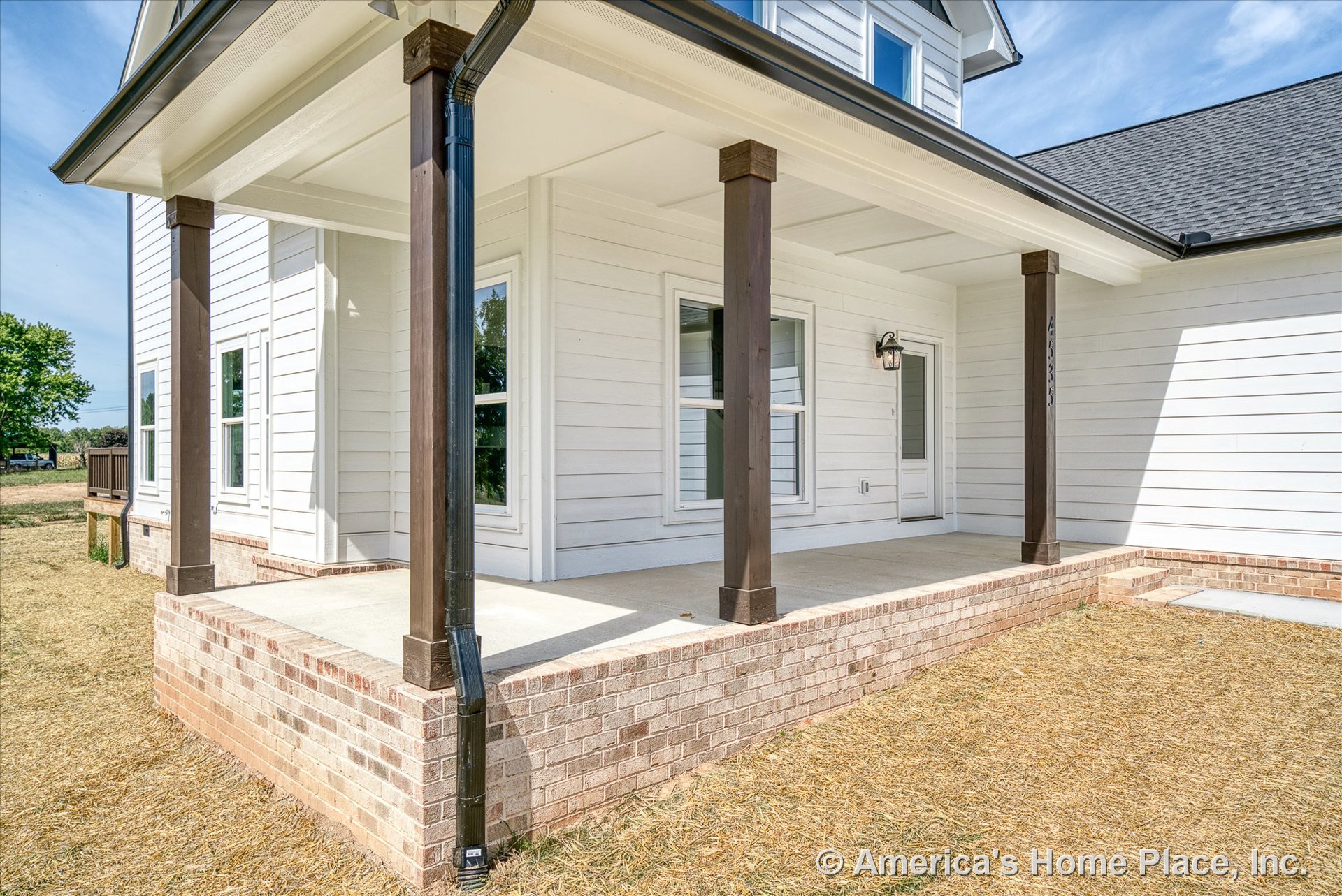 Covered front porch with white horizontal siding, dark wood columns, brick steps and base, concrete floor, double front windows, exterior wall-mounted light, and trim detailing