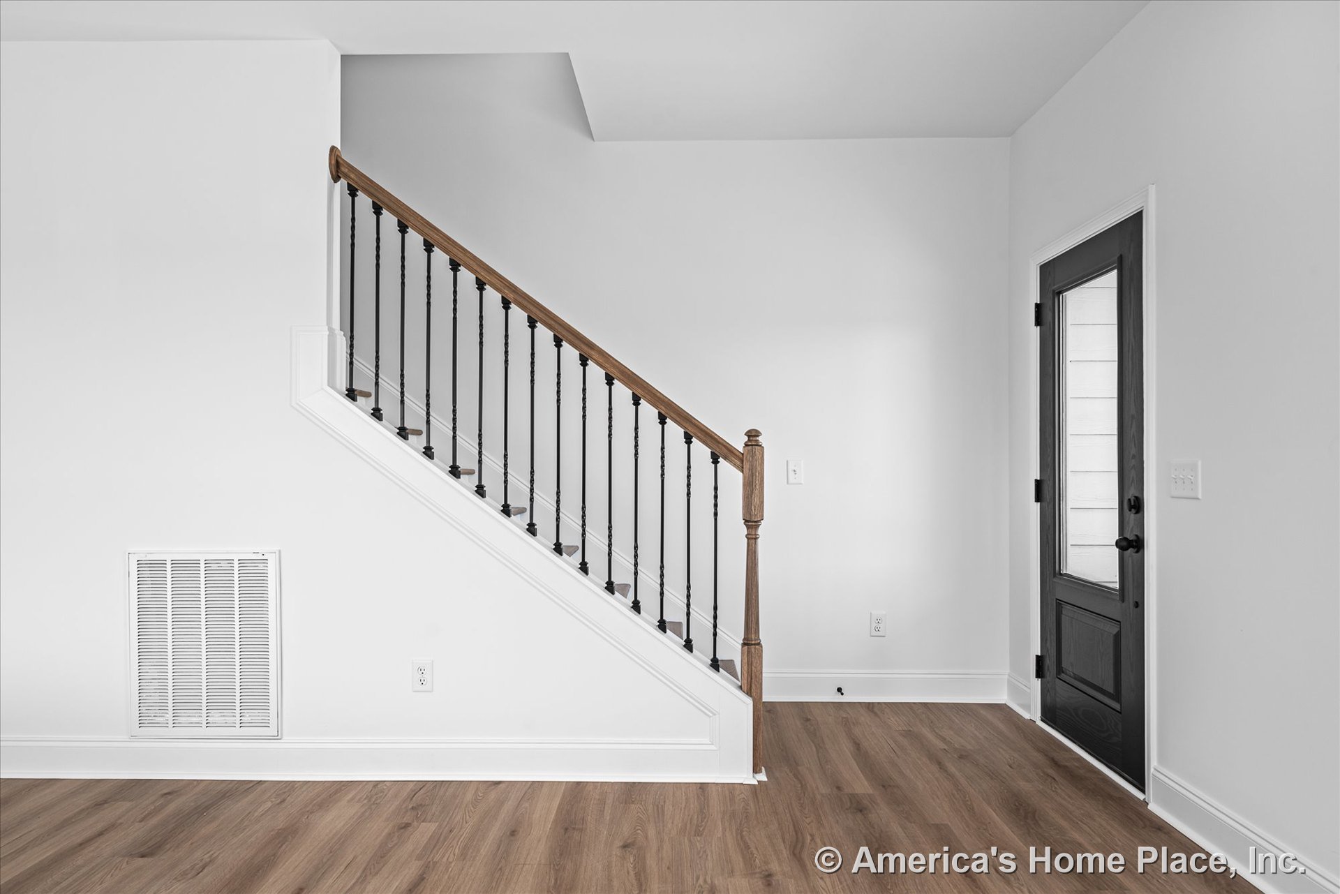 Staircase with wood handrail and black metal balusters beside a black framed glass door, white trim and baseboards, wall vent cover, and light wood flooring in the entryway.