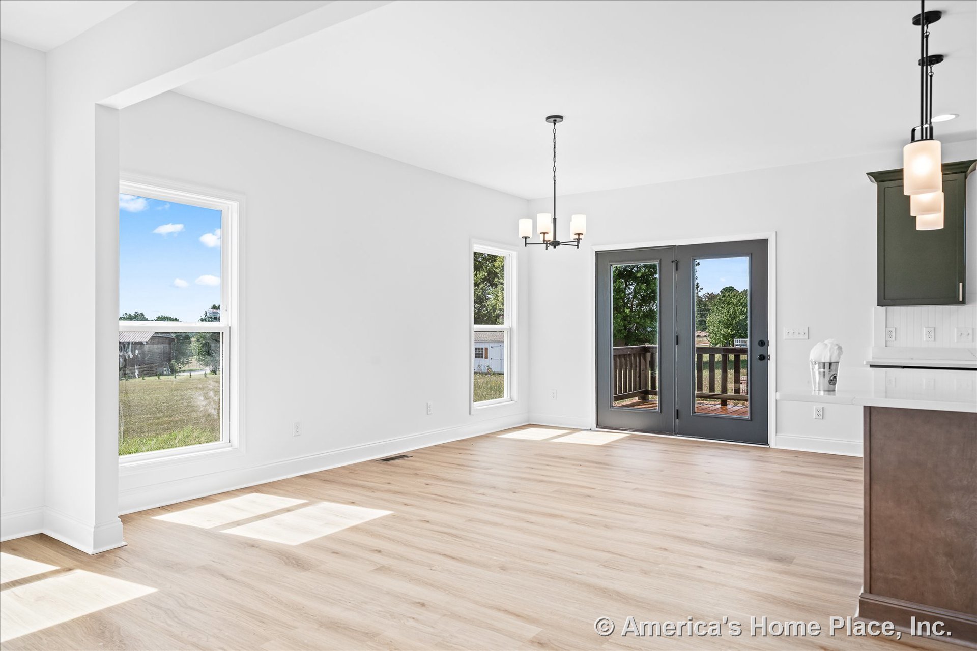 Dining area with light wood plank flooring, white walls and trim, large rectangular windows, double glass doors opening to porch, modern chandelier, and adjacent kitchen peninsula