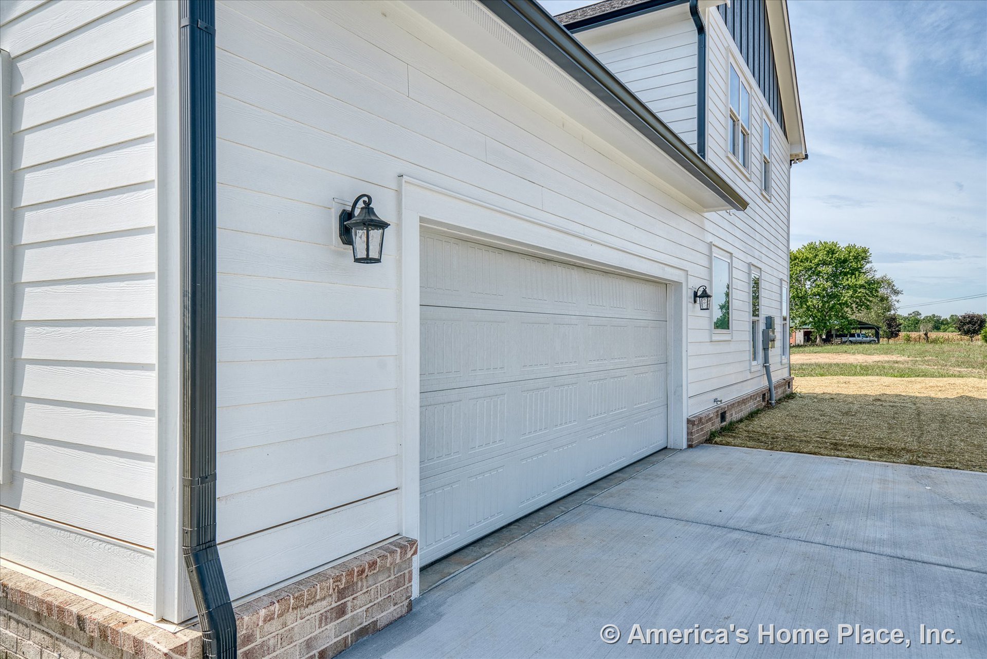 White horizontal siding exterior with double white paneled garage door, brick foundation trim, black gutter and downspout, wall-mounted lantern lights, and concrete driveway.
