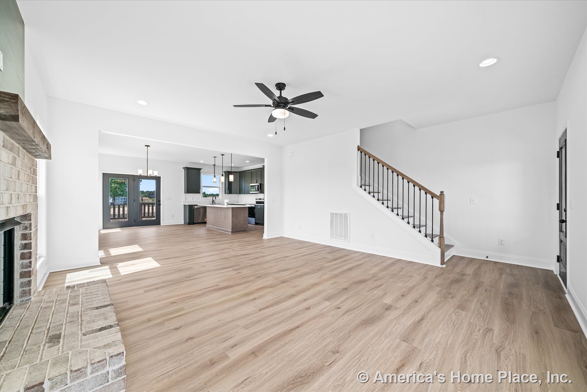 Open floor plan with light wood plank flooring, brick fireplace with wood mantel, kitchen featuring dark cabinetry and pendant lights over island, double glass doors leading to