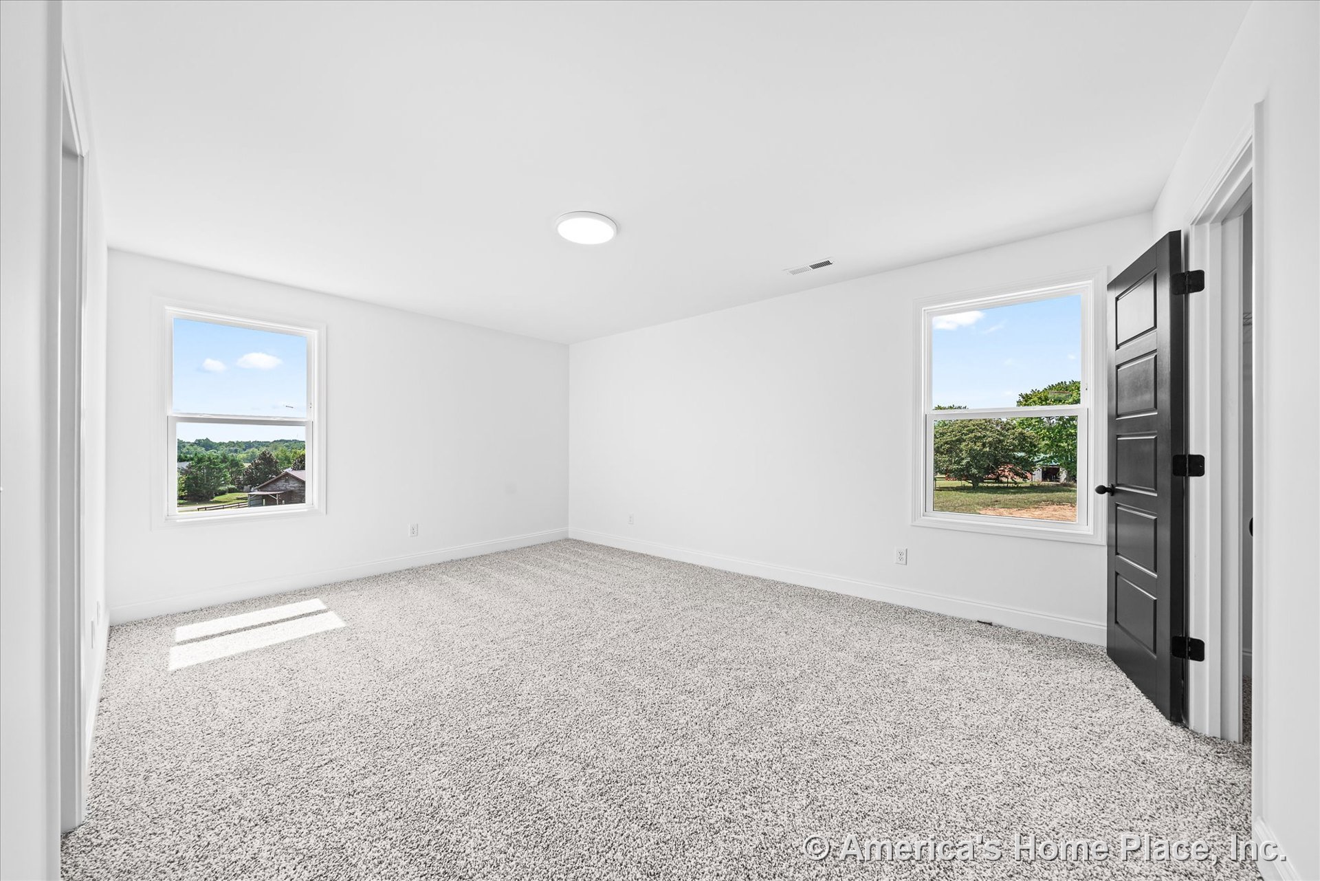 Bedroom with double large windows, white walls and trim, black paneled door, flush ceiling light, neutral plush carpet flooring, smooth white ceiling, and baseboards.