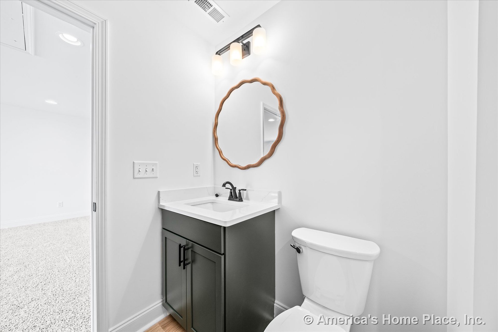 Dark wood bathroom vanity with white quartz countertop, round wall mirror, three-light fixture above, white toilet, and light wood flooring.