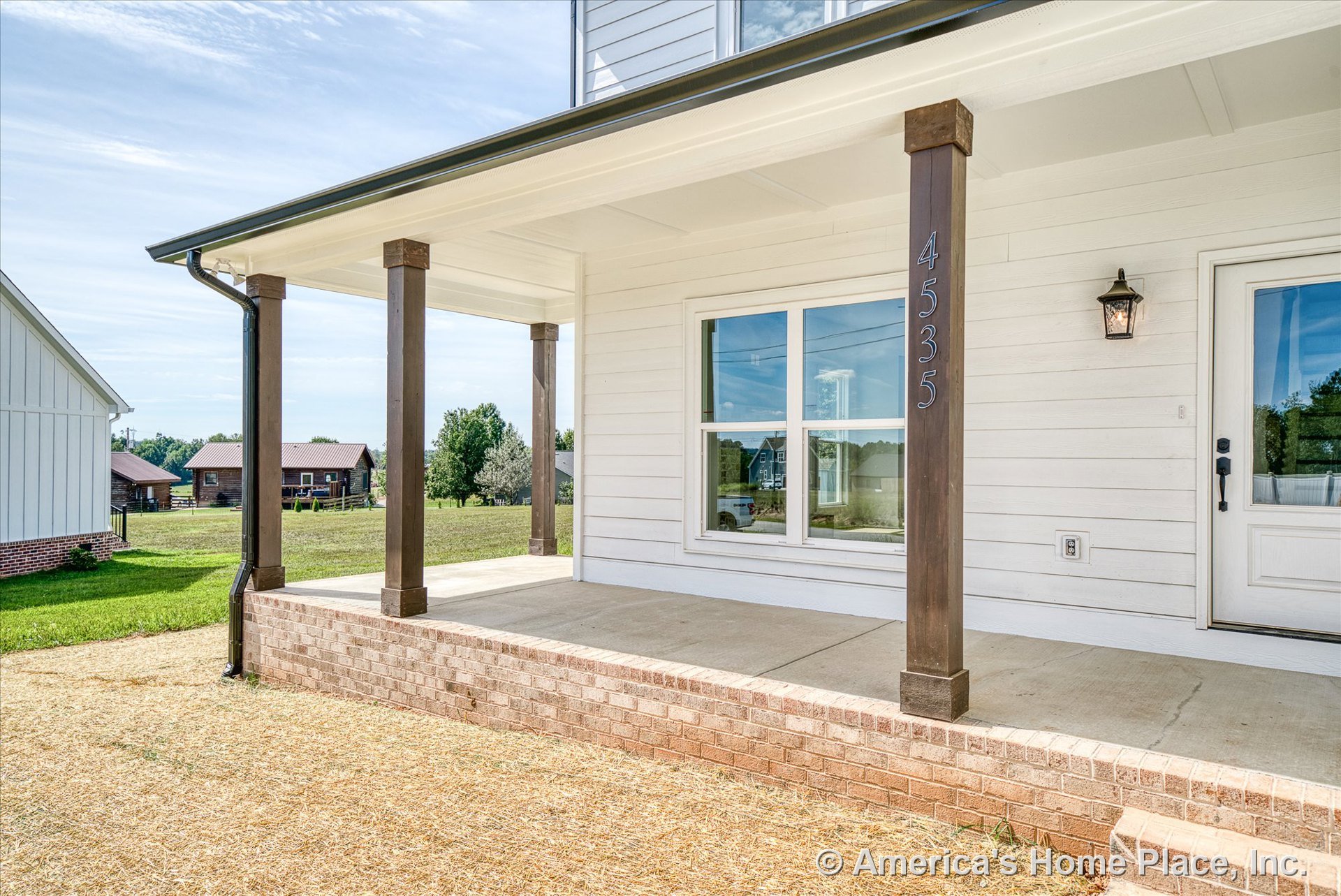 Covered front porch with brick steps, concrete floor, wood support columns, white horizontal siding, large double window, exterior wall lantern, and decorative trim near the entry