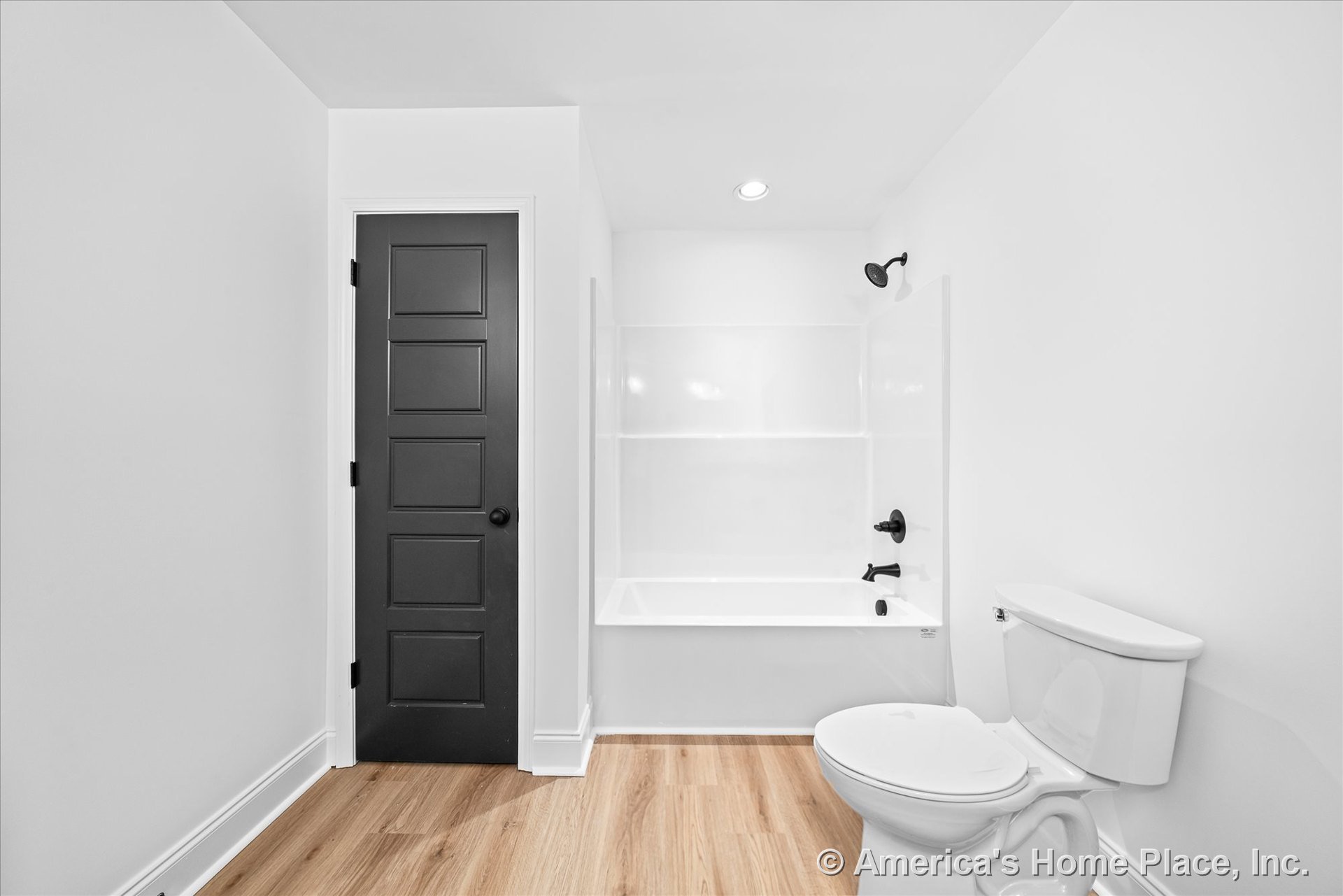 White bathtub with integrated shower, matte black faucet and showerhead, light wood plank flooring, white toilet fixture, black panel door, recessed ceiling light, white walls, and