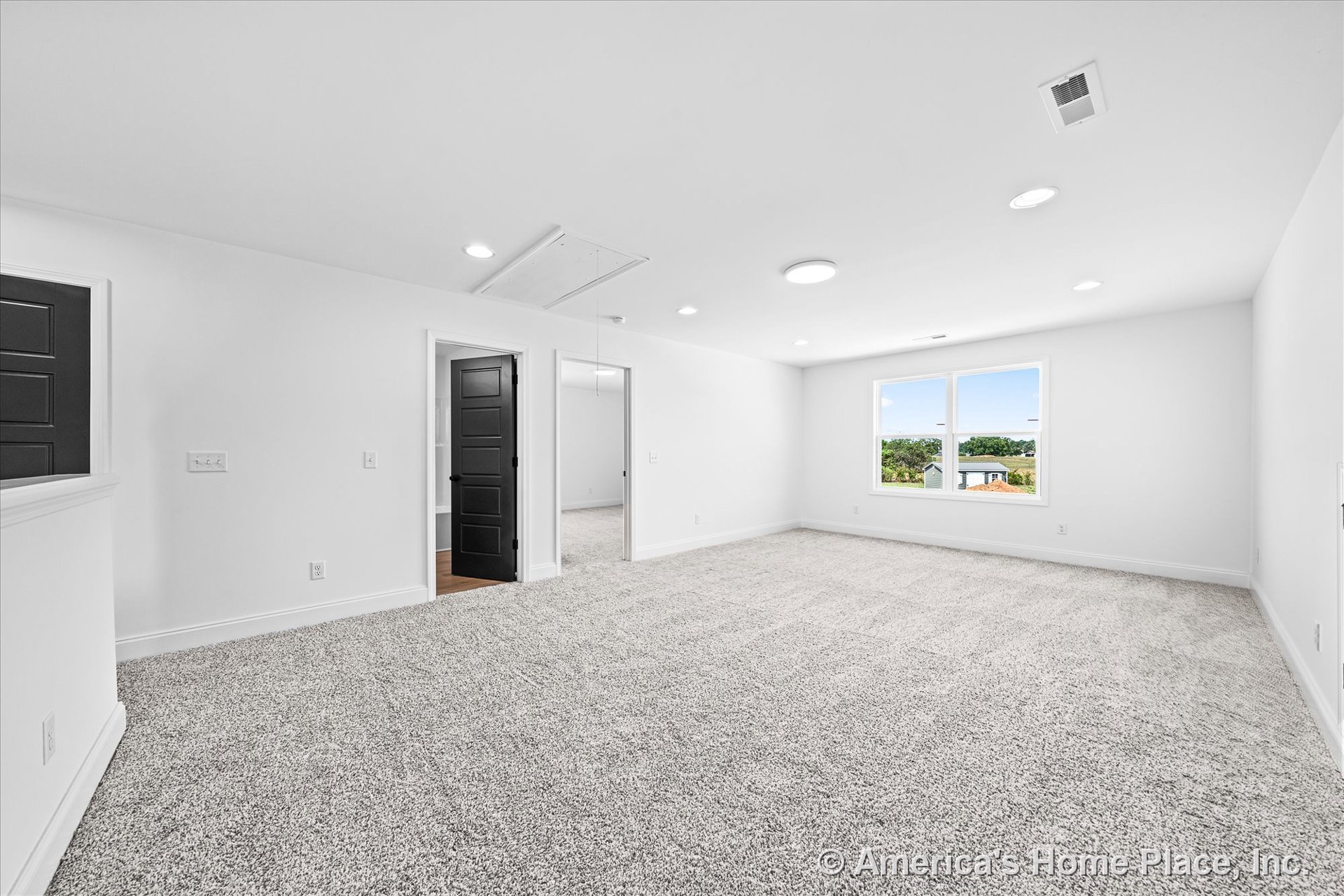 Spacious upstairs bonus room with light gray carpet, recessed ceiling lights, large double window framed in white trim, white walls and baseboards, black paneled doors, attic