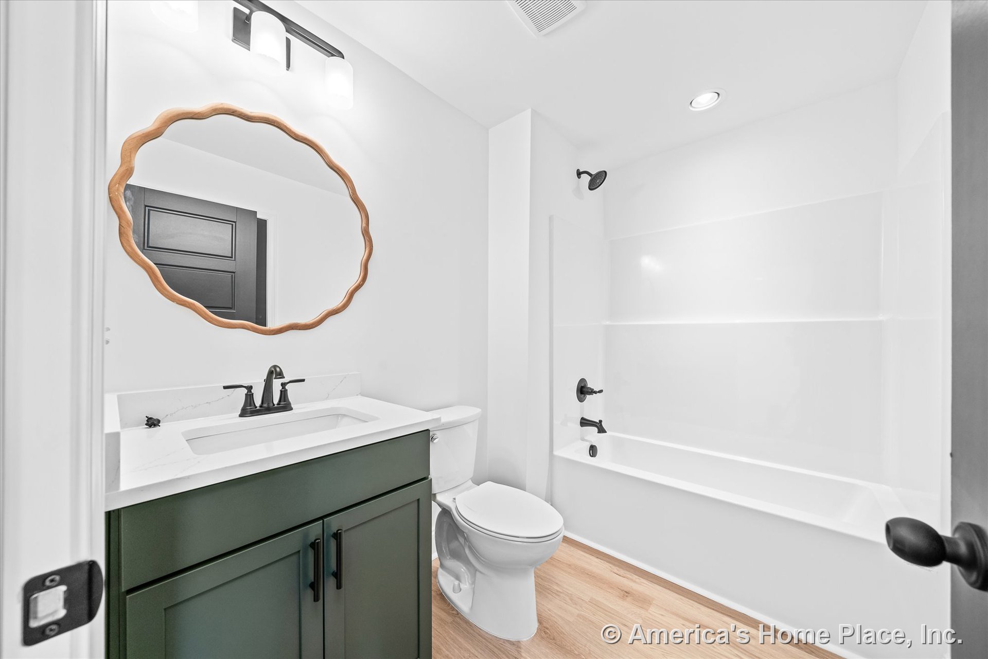 Green vanity with white quartz countertop, scalloped wood-framed mirror above, built-in bathtub and shower with matte black fixtures, light wood flooring, white walls, recessed