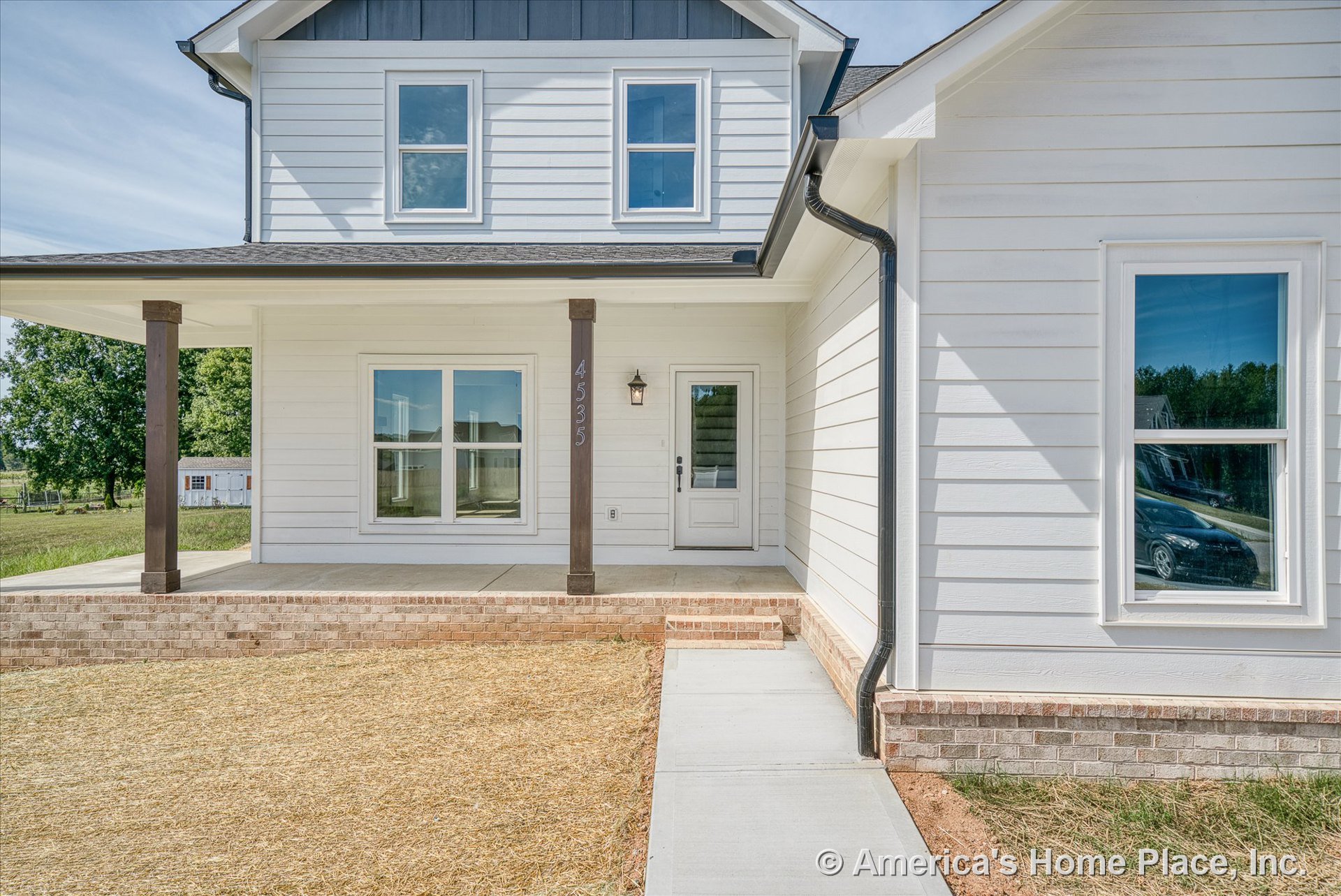Covered front porch with brick steps, white horizontal siding, double front windows, dark wood columns, exterior wall lantern, concrete walkway, and visible house number.