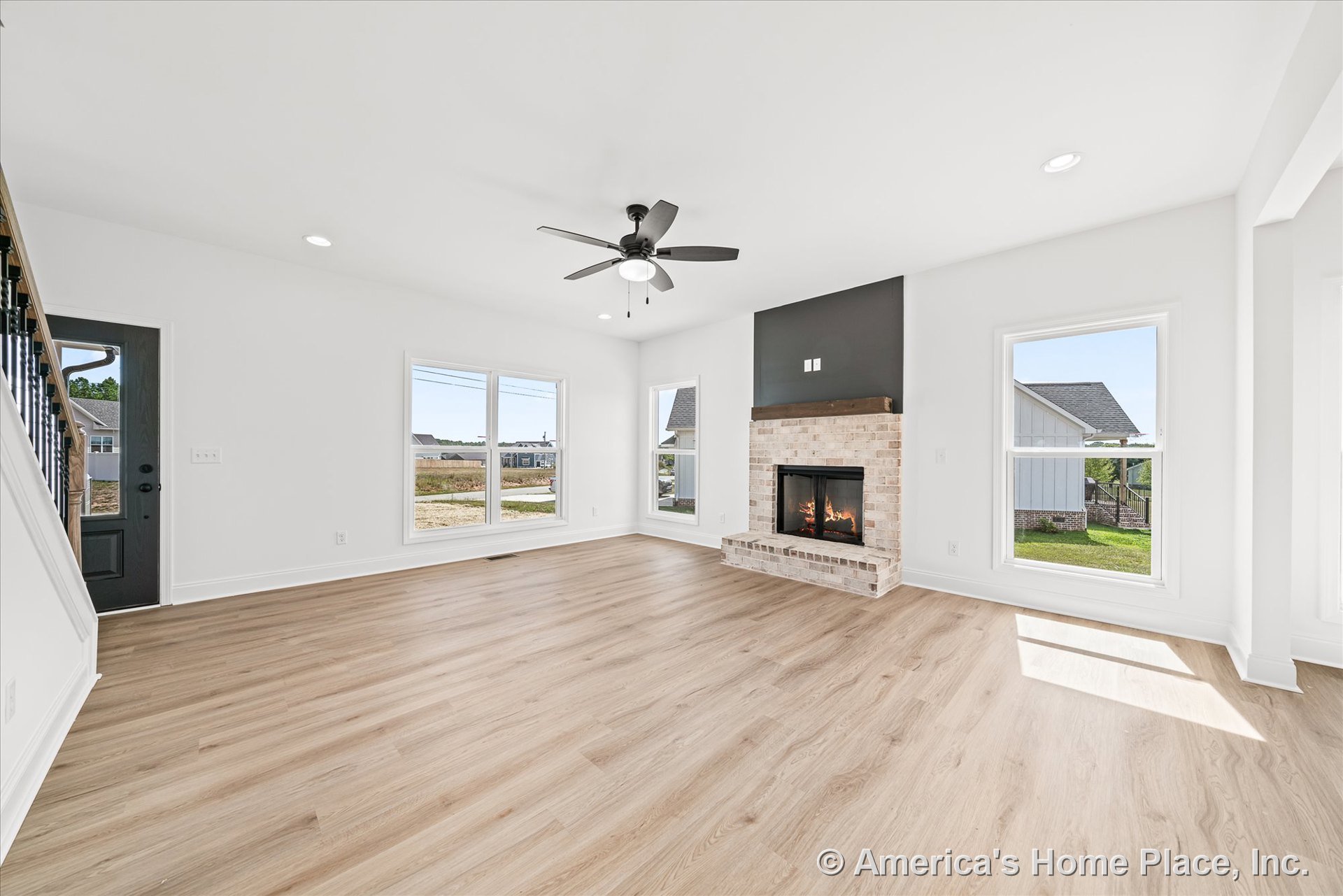 Brick fireplace set against a dark accent wall, light wood plank flooring, large double windows with white trim and baseboards, black ceiling fan with light fixture, black entry