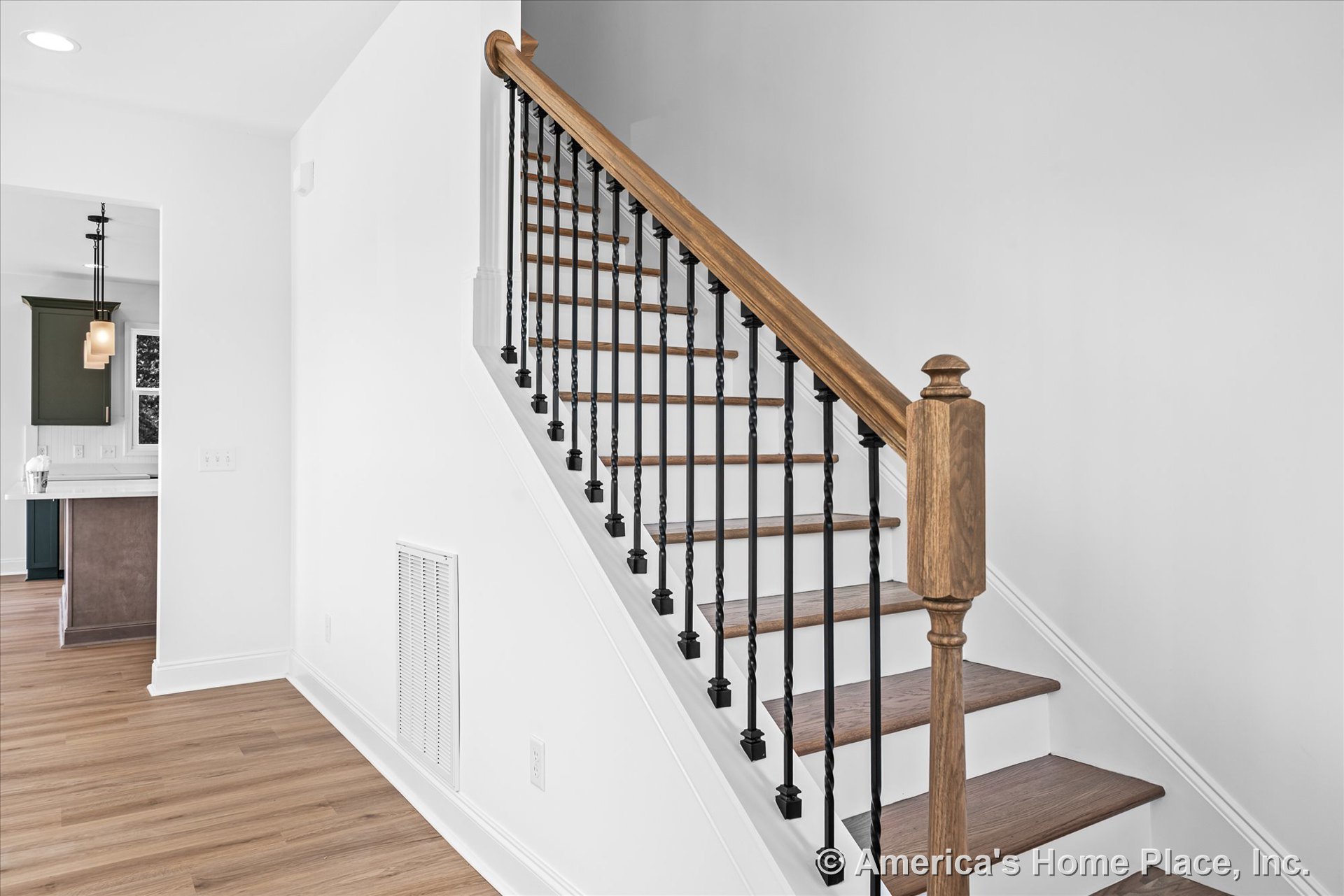 Staircase with wood treads, white risers, black metal balusters, and wood handrail; open entry to kitchen; light wood flooring; white walls and trim; ceiling lighting fixture and