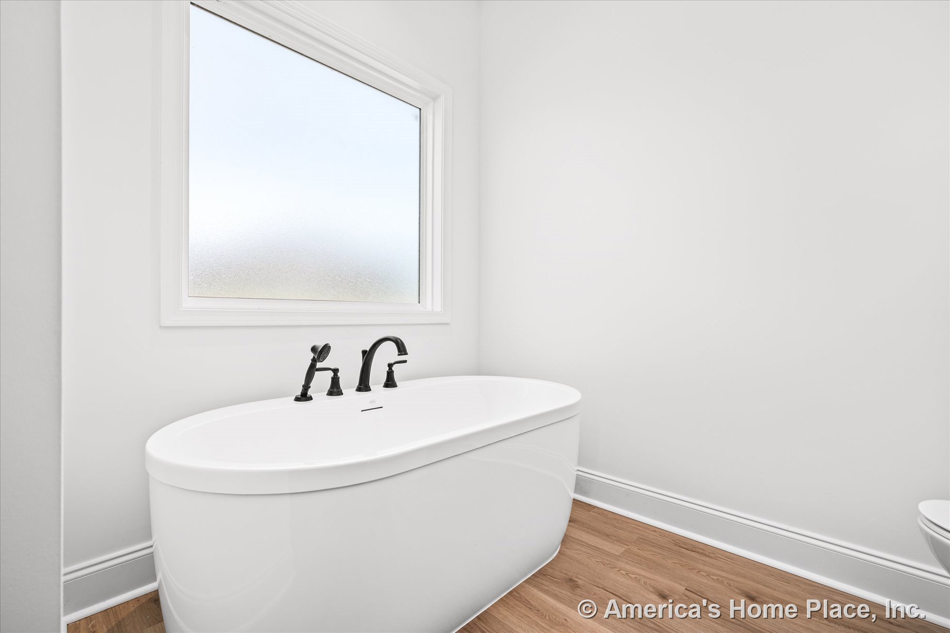 Freestanding soaking tub with matte black faucet beneath frosted window, white trim, light wood flooring, and white walls in a modern minimalist bathroom.