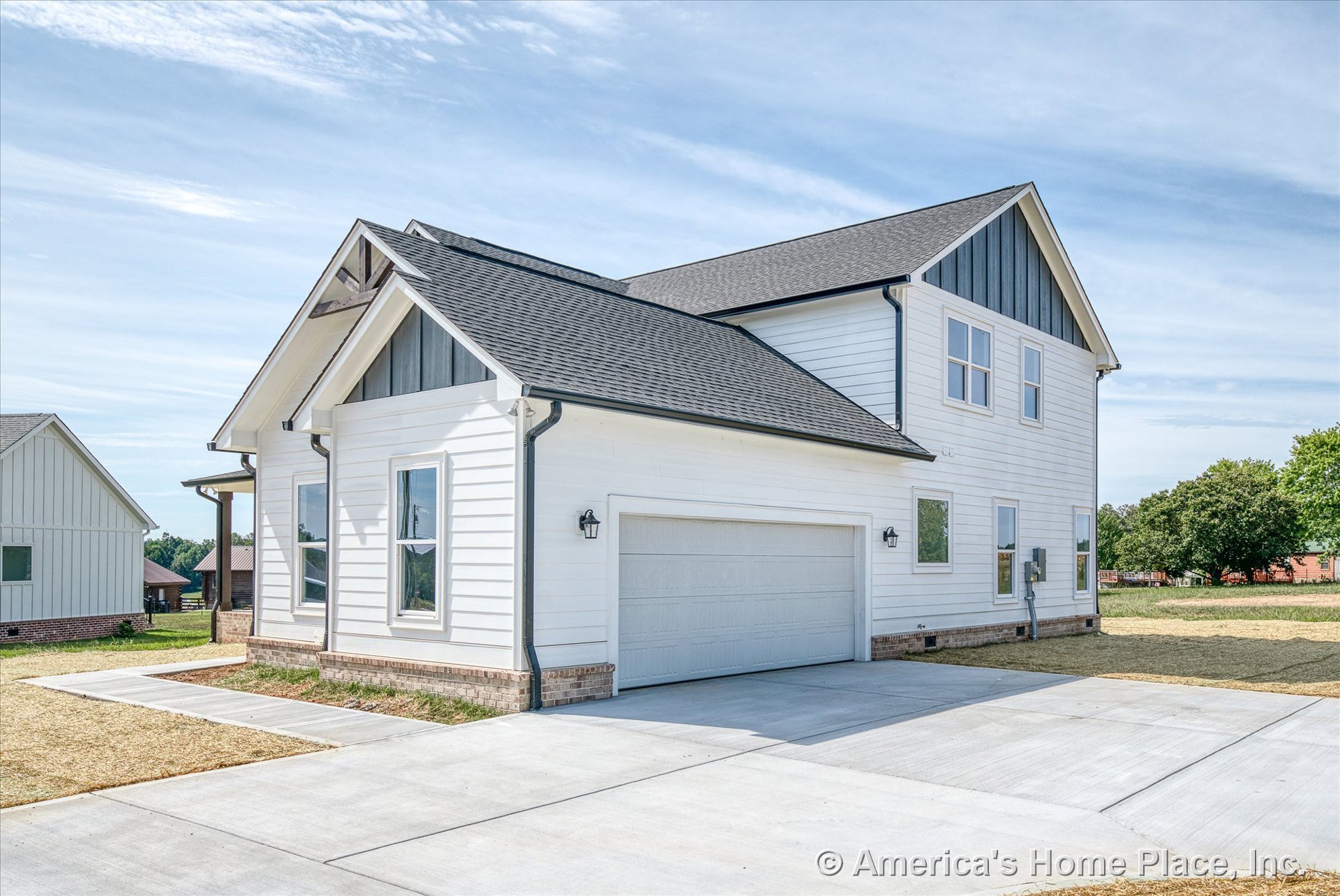 White horizontal siding with brick foundation trim, two-car garage door, architectural gable accents, black roof shingles, exterior wall-mounted lights, concrete driveway, covered