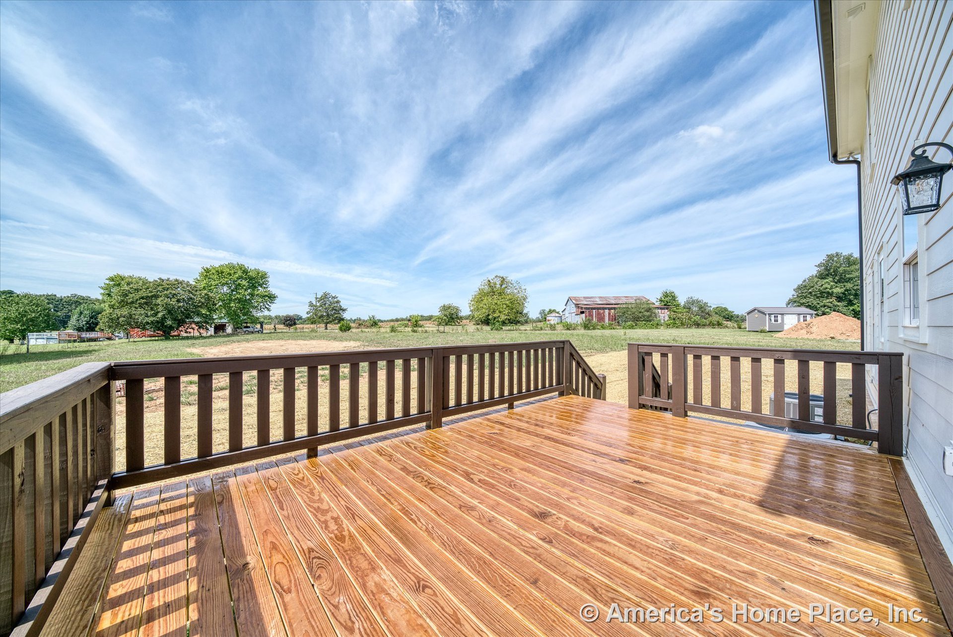 Freshly stained wooden deck with horizontal plank flooring, vertical slat brown railing, exterior wall siding, outdoor lantern light fixture, and steps overlooking an open rural
