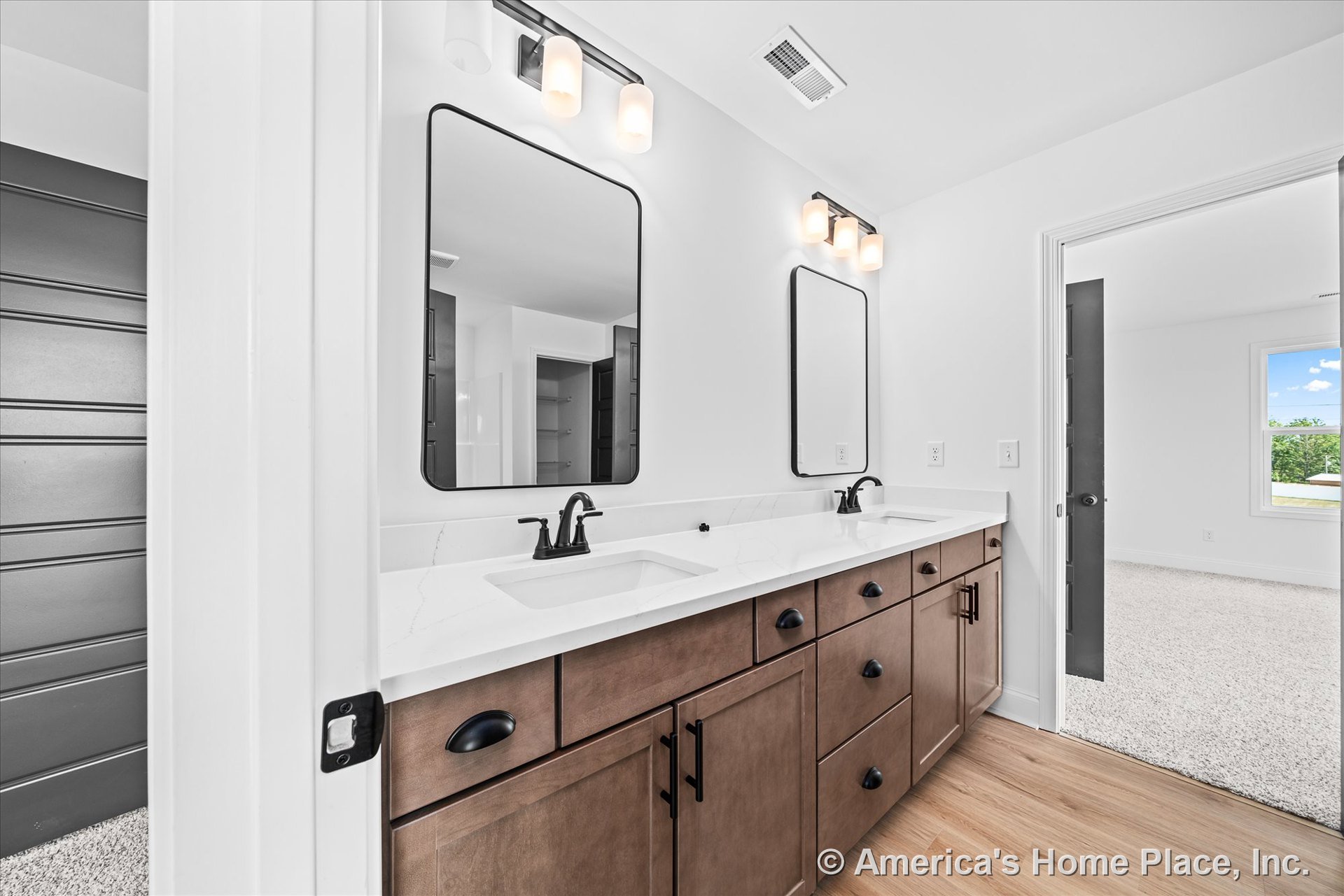 Double vanity with wood cabinetry, quartz countertops, black faucets, rectangular mirrors, built-in vanity lighting, white walls, modern trim, and light wood flooring.