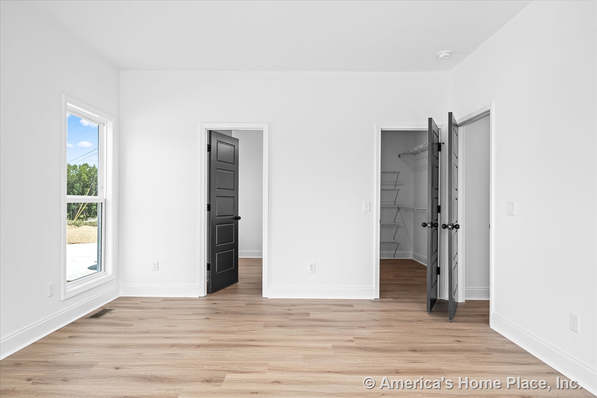 Bedroom with light wood plank flooring, smooth white walls, black paneled interior doors, large window framed in white trim, walk-in closet featuring wire shelving, white