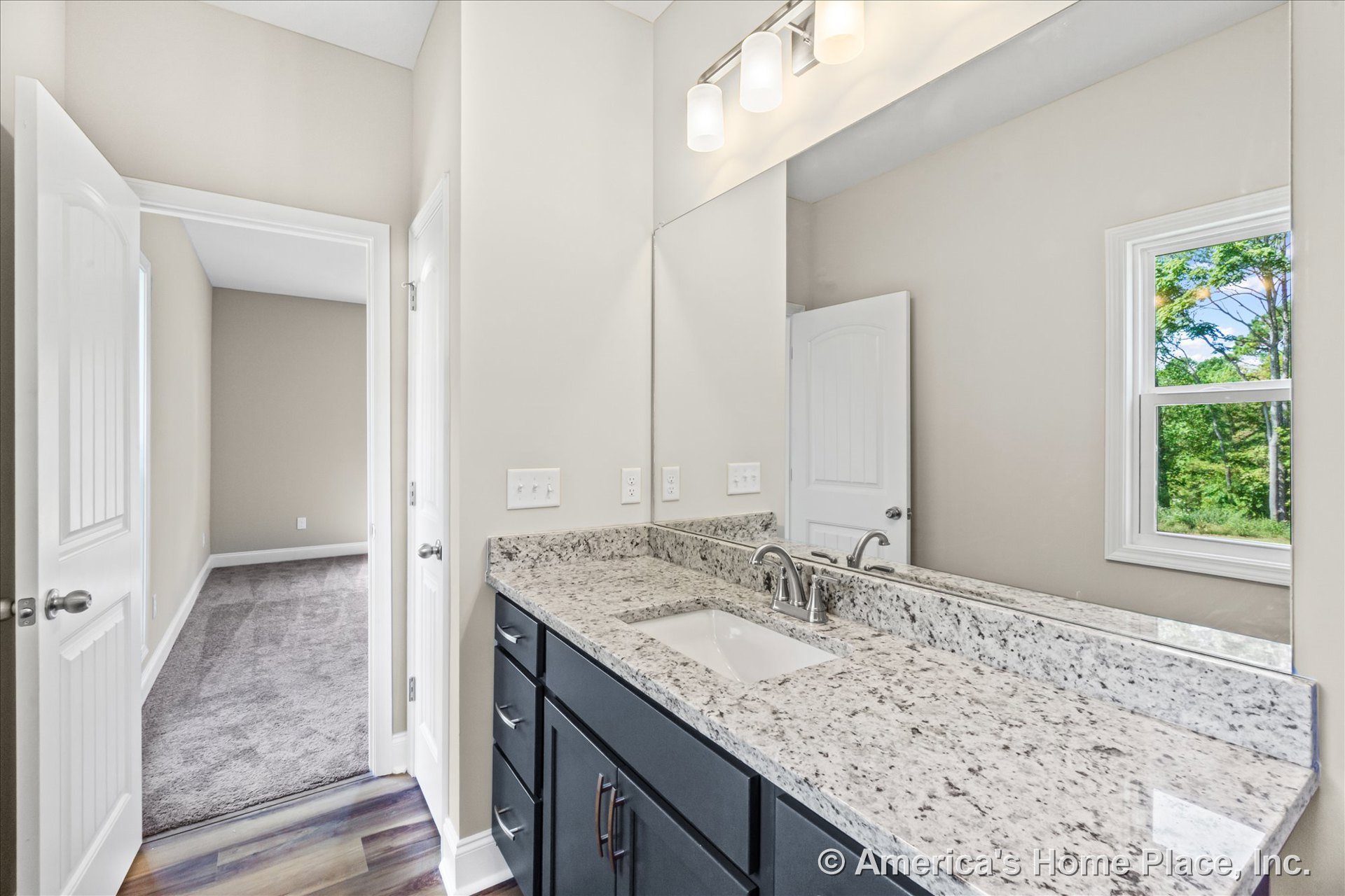 Granite vanity with undermount sink and dark wood cabinets beneath a large wall mirror, three-light fixture above, white trim window, interior doorways, neutral walls, vinyl plank