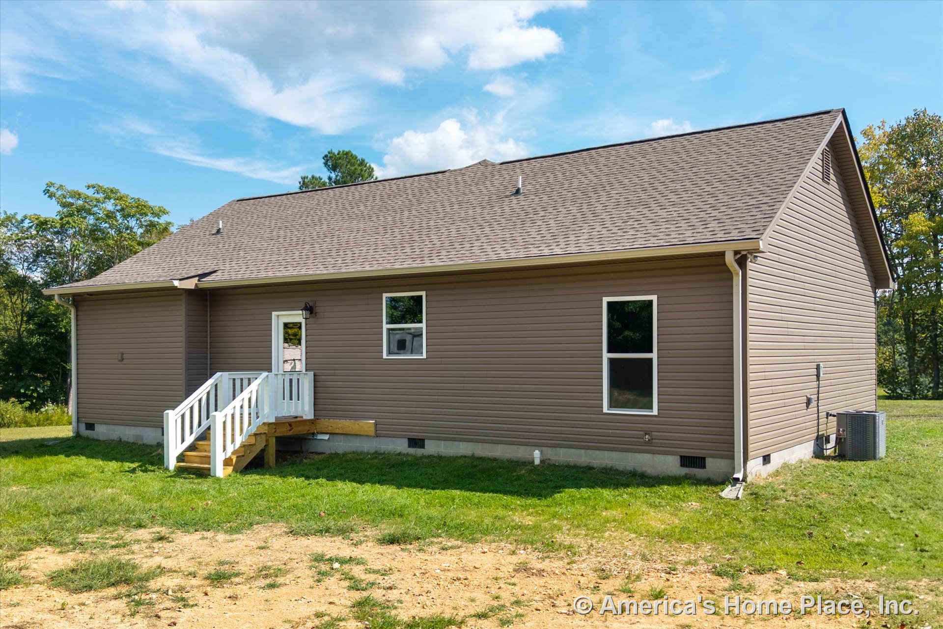 Tan vinyl siding and shingle roof with a small entry porch featuring white railings, two rectangular windows, exterior door with mounted light fixture, central air conditioning