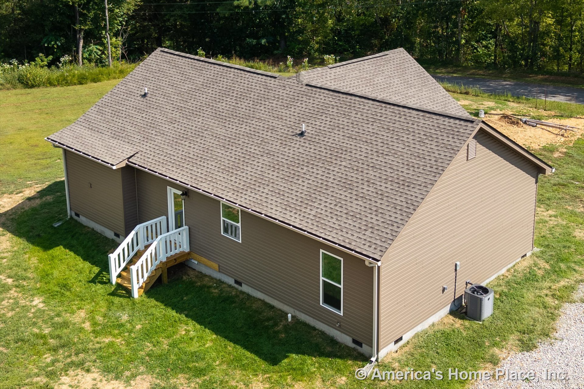 Tan vinyl siding exterior with asphalt shingle roof, small entry porch featuring white railings, rectangular windows, central HVAC unit beside concrete foundation, single-story