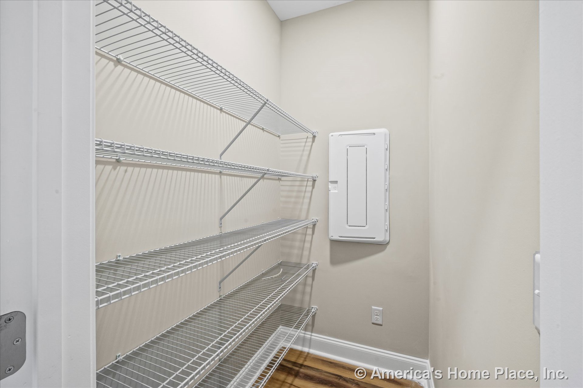 Built-in wire shelving units line a recessed pantry space with a wall-mounted electrical panel, light beige walls, wood-look flooring, and white baseboard trim.
