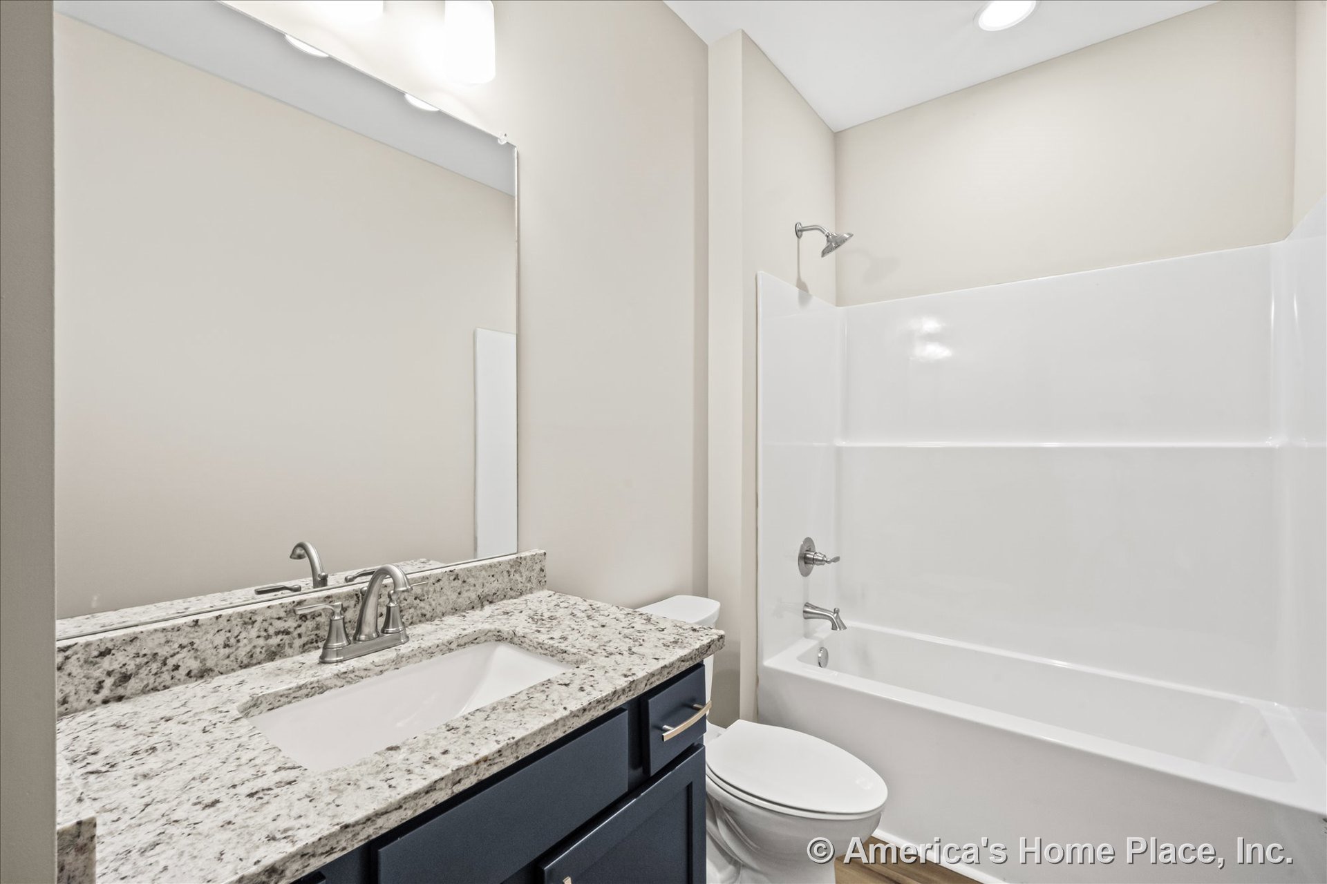 Granite countertop vanity with single undermount sink, dark cabinetry and modern hardware, built-in bathtub with shower surround, large wall mirror, recessed ceiling light