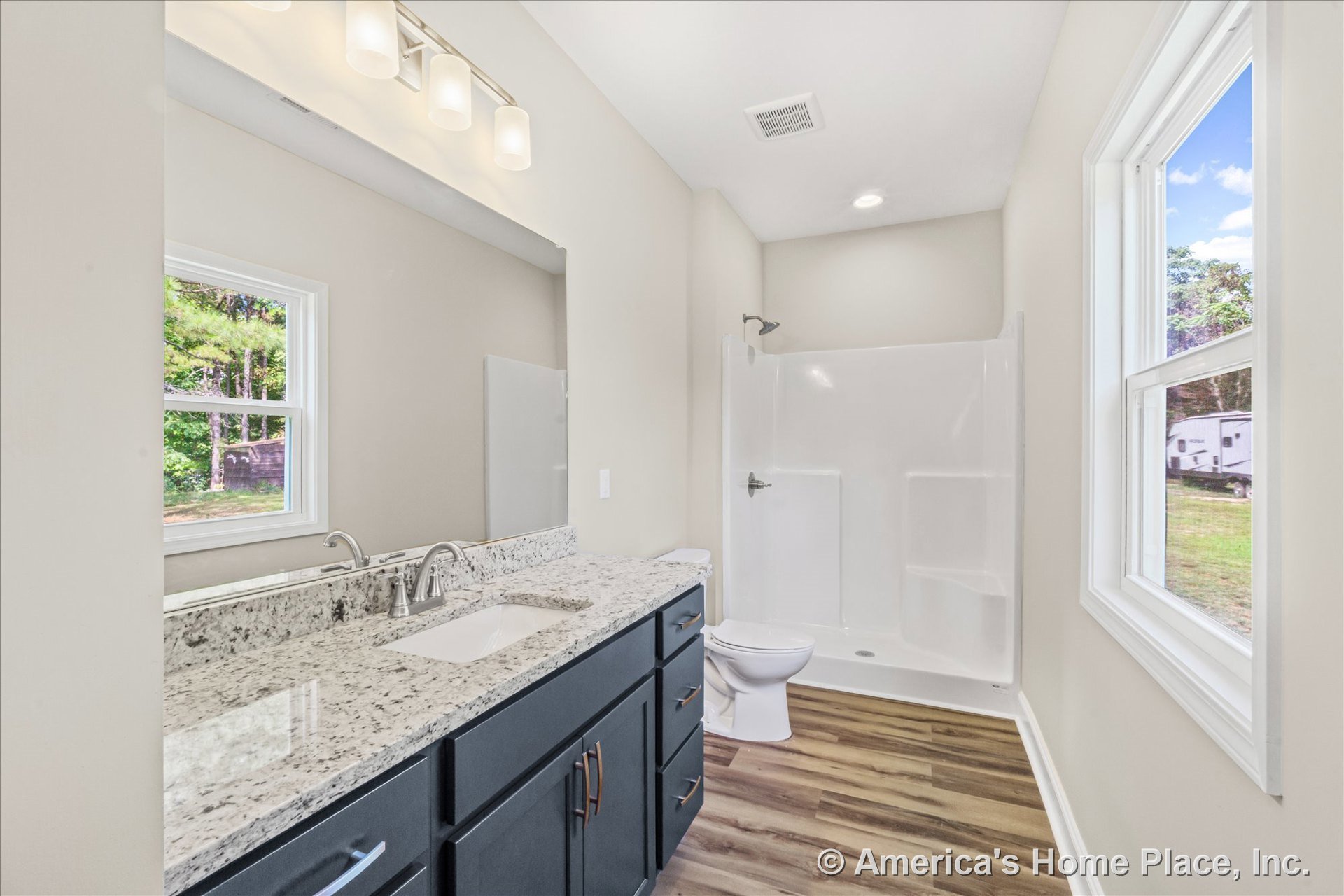 Granite countertop vanity with undermount sink and dark cabinetry featuring modern hardware, walk-in fiberglass shower enclosure, wood-look vinyl plank flooring, large wall mirror
