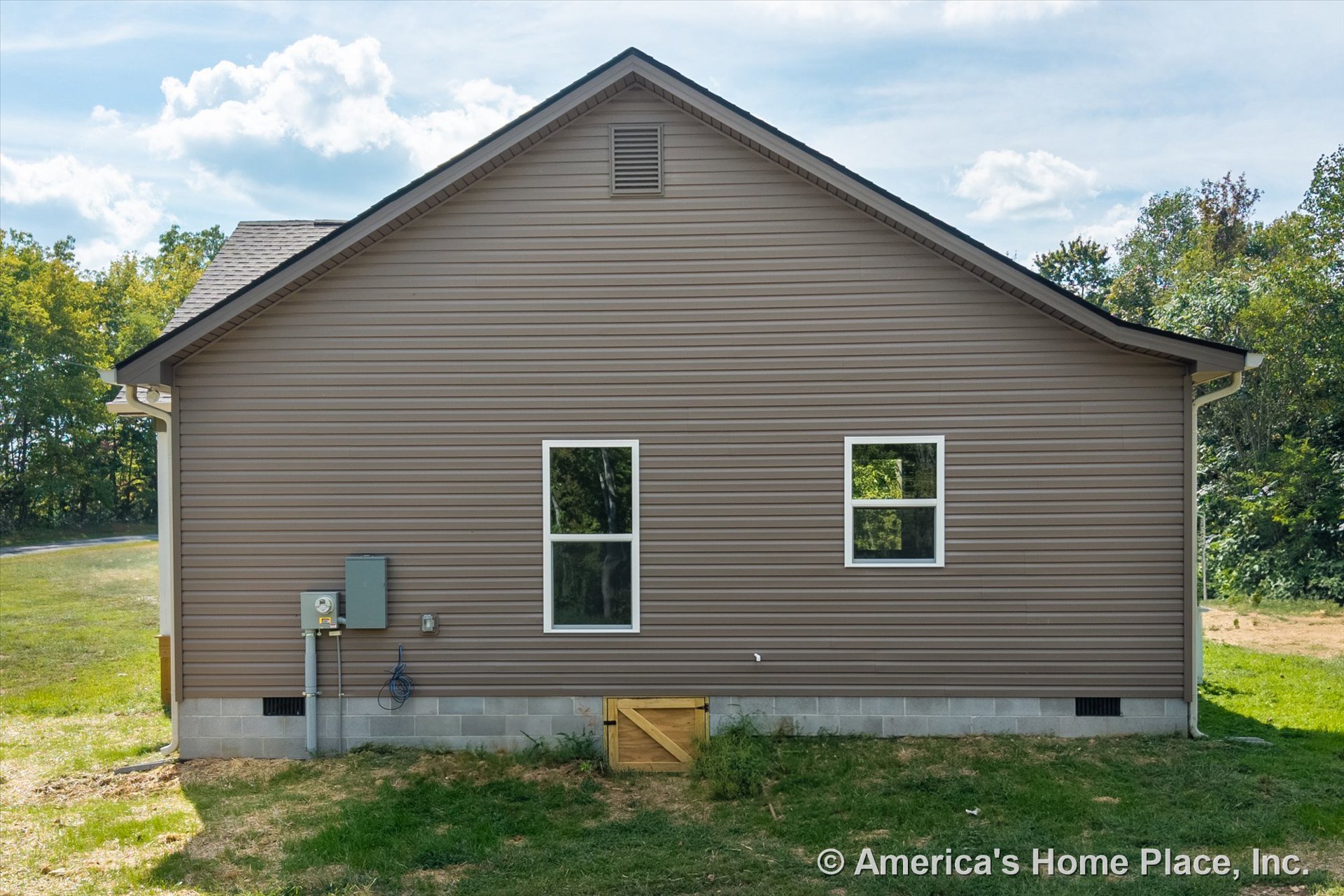 Brown vinyl siding exterior with two rectangular windows, vented gable, concrete block foundation, utility meter and electrical panel, crawl space access door, and white trim.